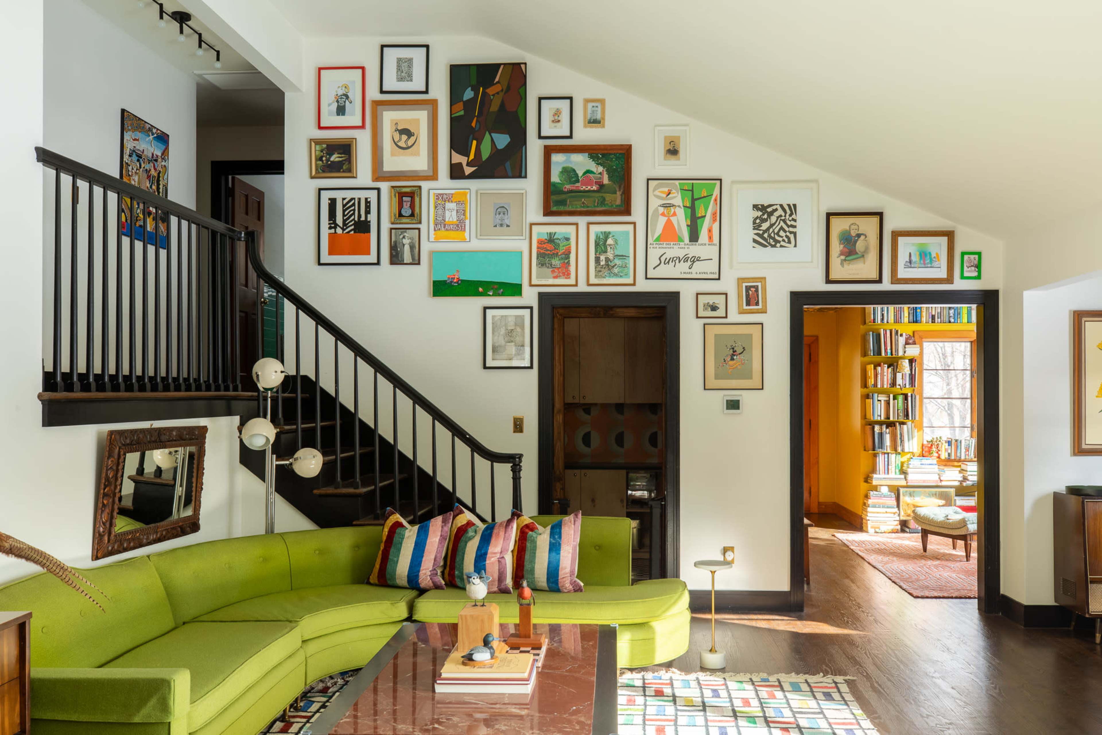 The image shows a bright and eclectic living room with a green sectional sofa, a gallery wall of framed art, and a staircase leading to an upper level.