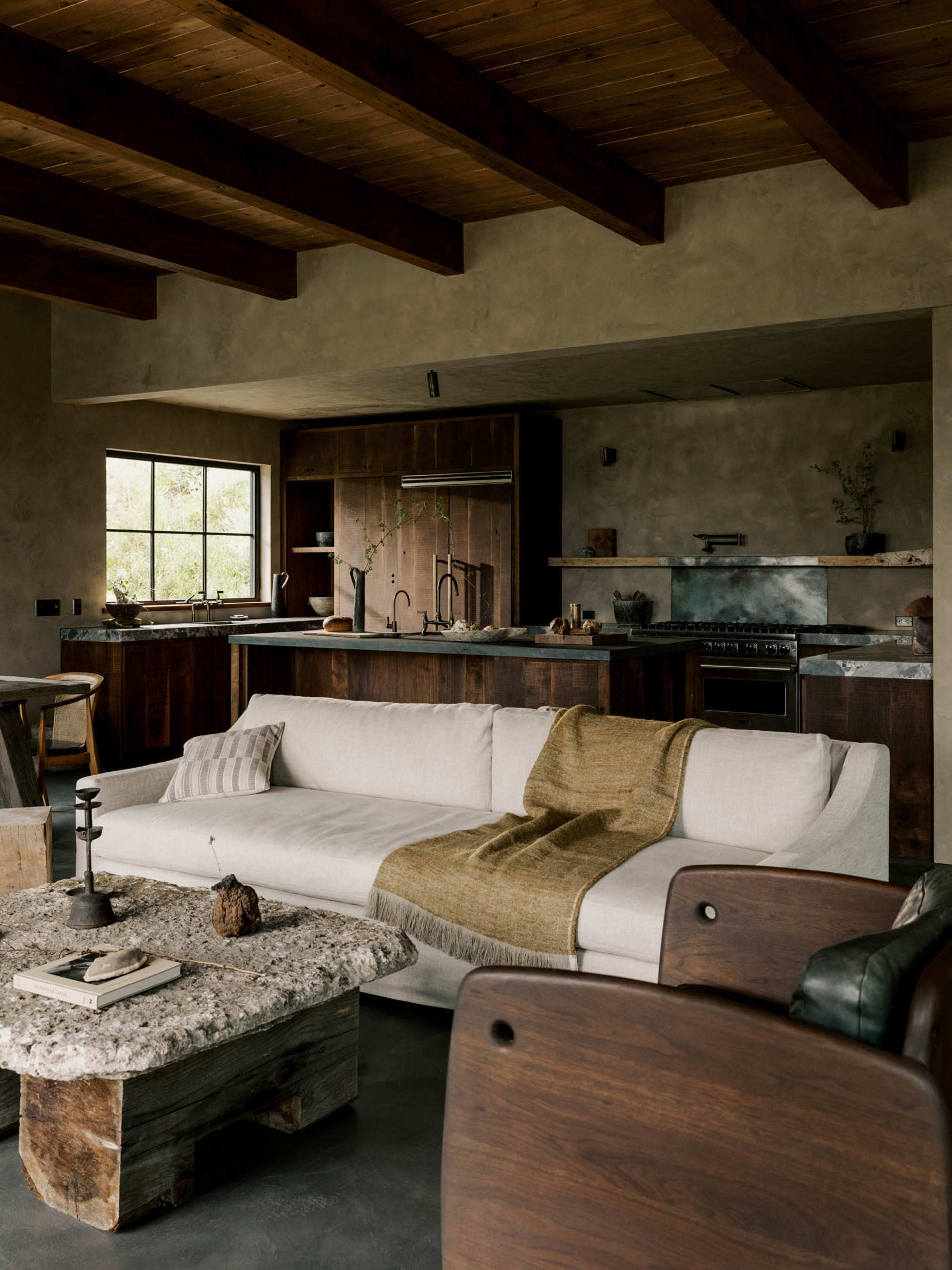 The image shows a modern living room with a light-colored sofa, a rustic coffee table, and an open kitchen featuring wooden cabinetry.