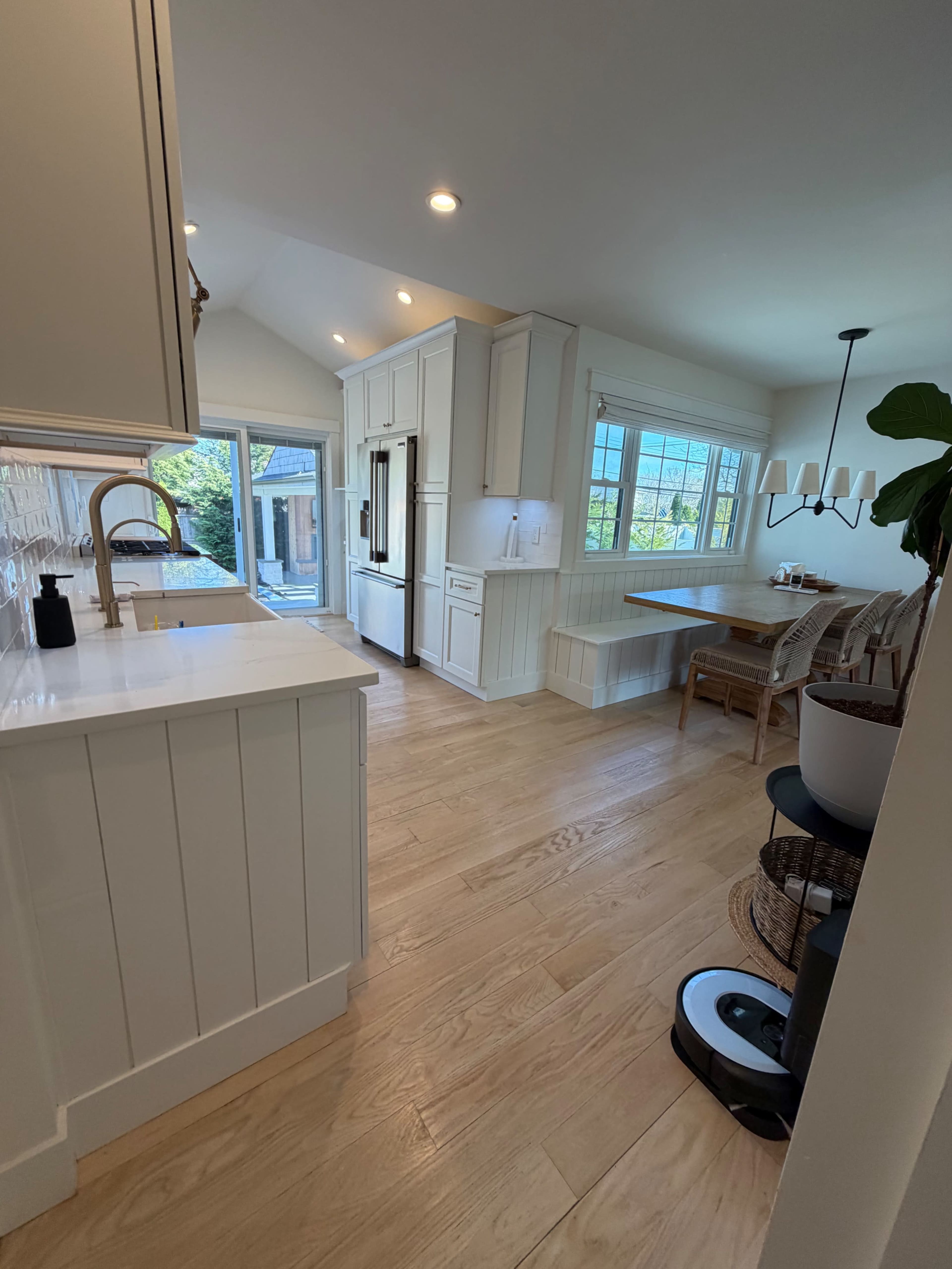 The image depicts a modern kitchen with white cabinetry, a wooden dining table, and a large window allowing natural light to enter the space.