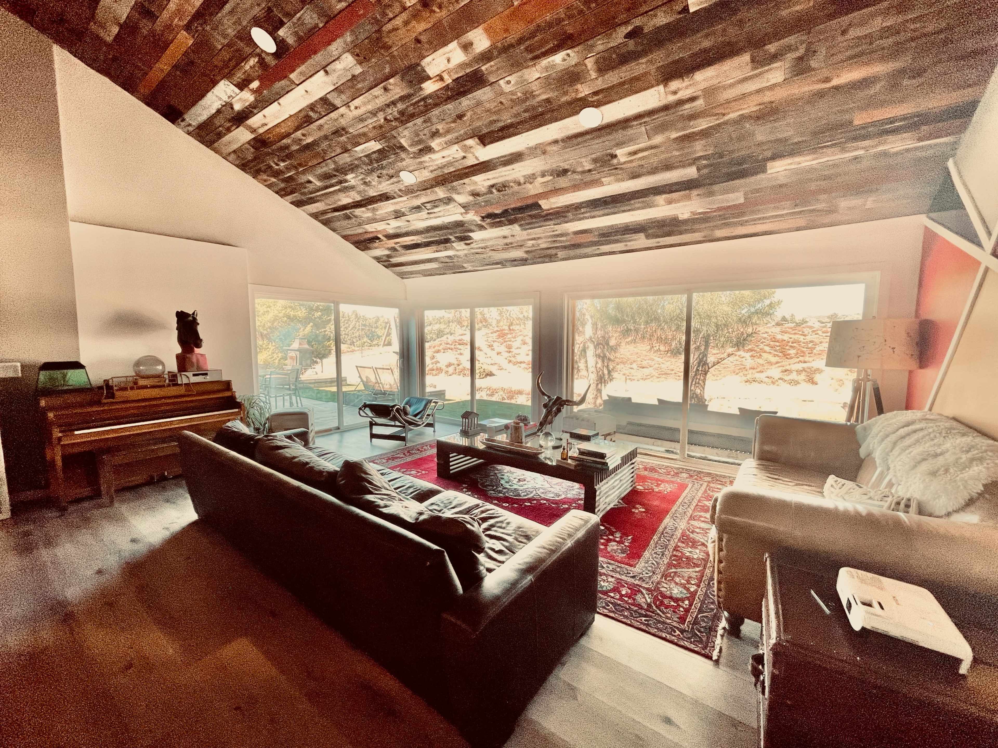 A spacious living room with a wooden ceiling, large windows overlooking a natural landscape, and furniture including sofas and a piano.