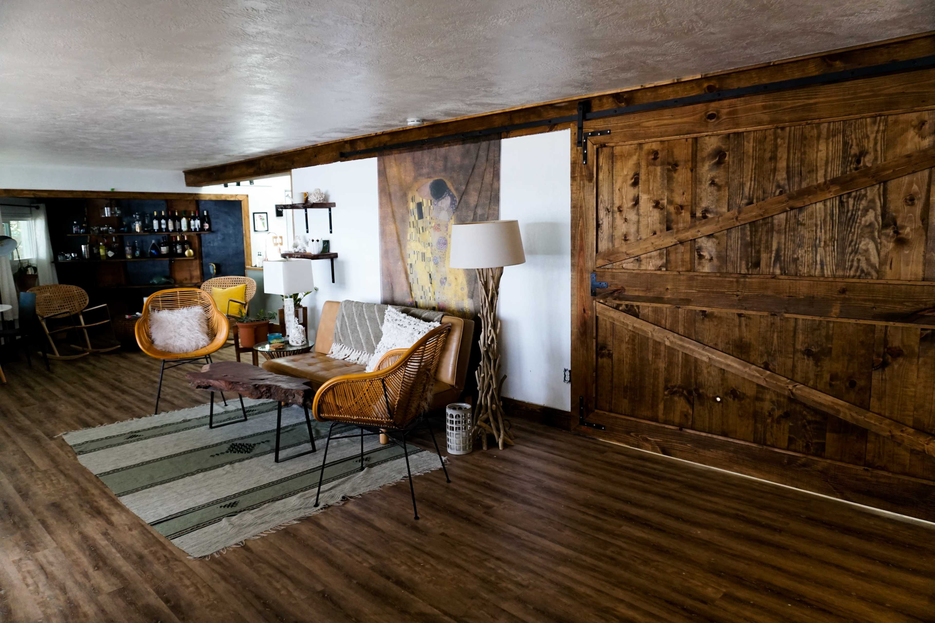 The image shows a sitting area featuring a wooden barn-style door, a comfortable couch with wicker chairs, and a patterned rug on a wooden floor.