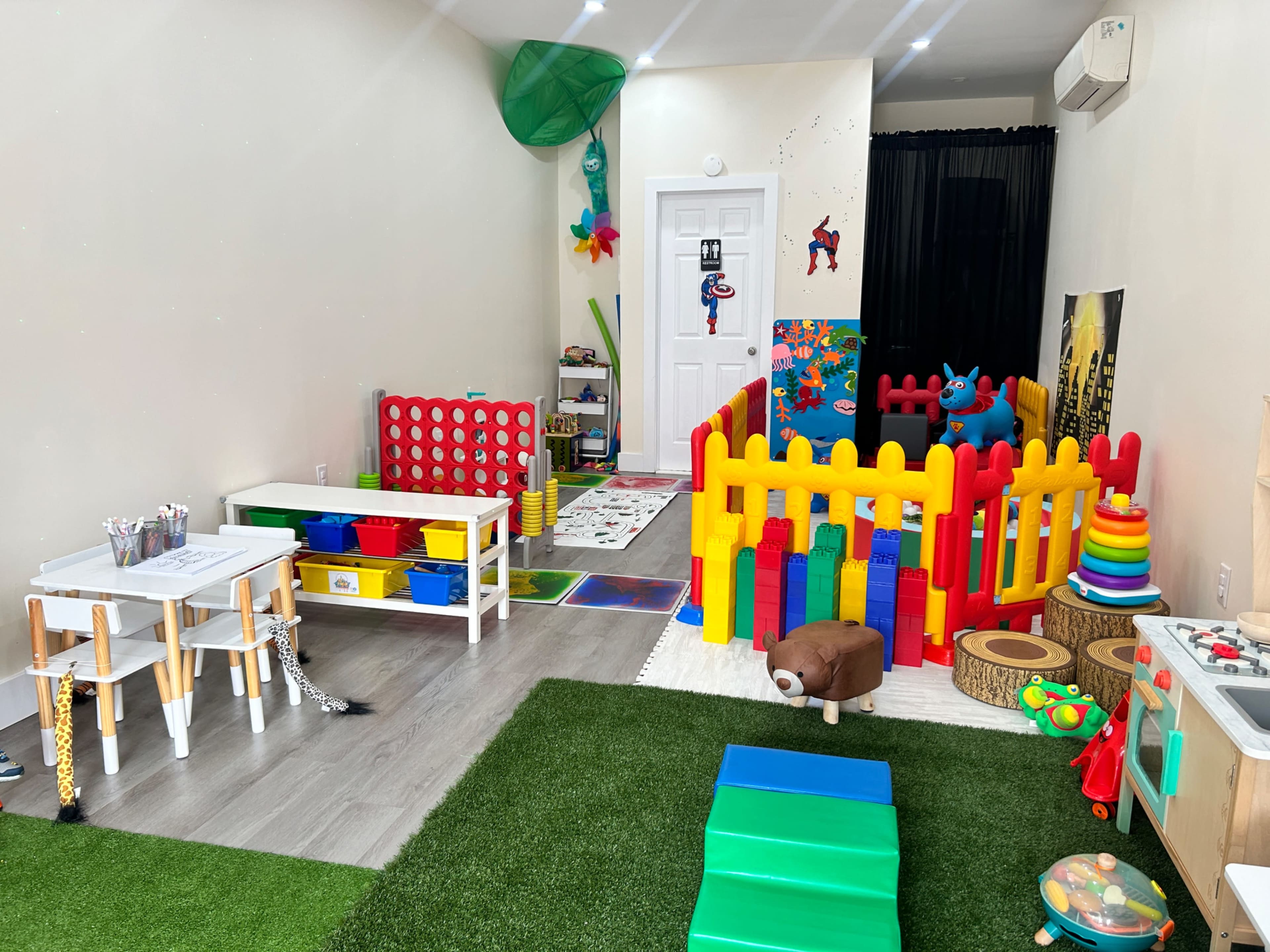 A colorful playroom filled with various children's toys, including a fenced play area, tables, and a green carpet.