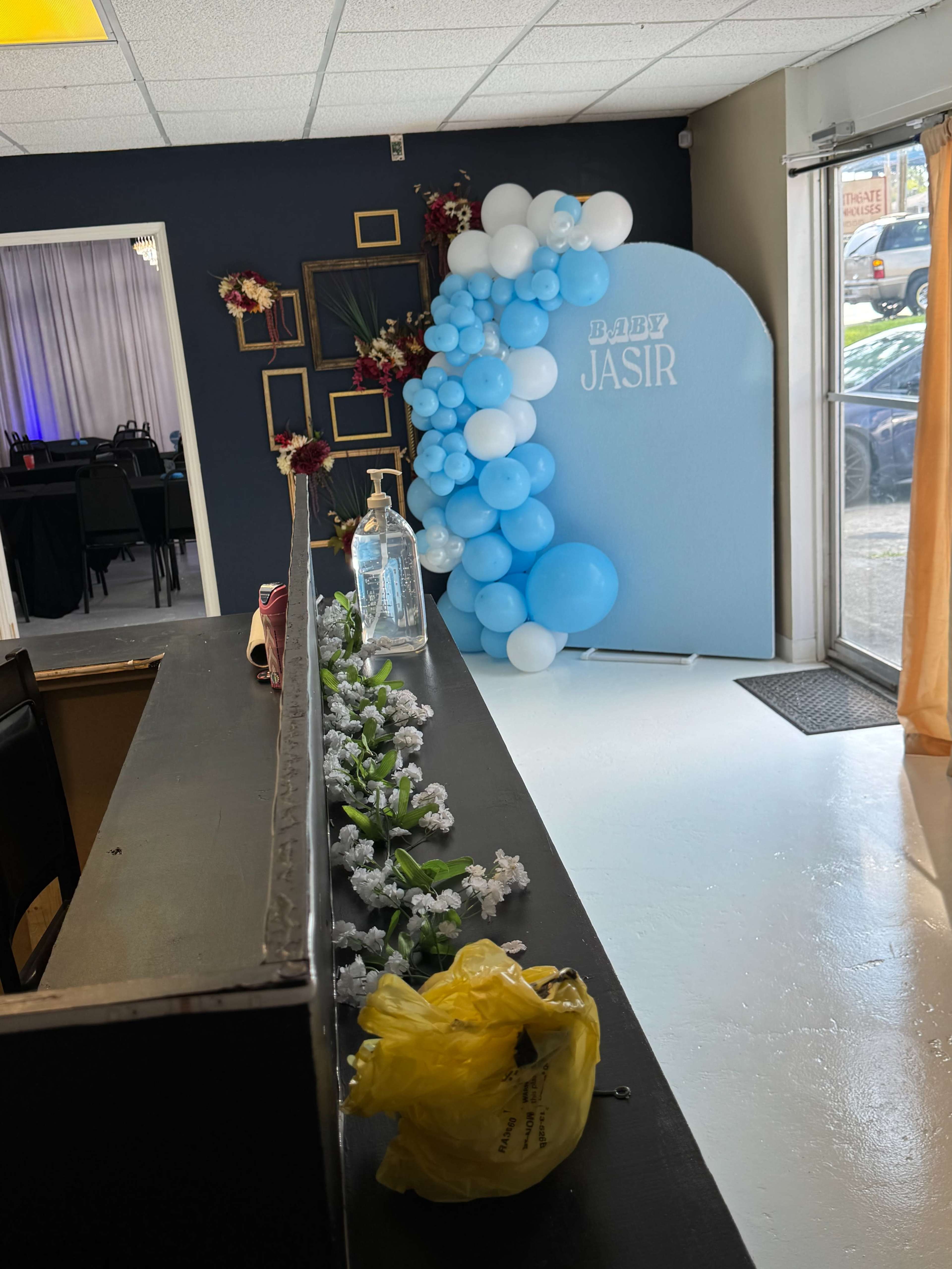 The image shows a decorated event space featuring a blue balloon arch with the text "Baby Jasir," a floral arrangement, and a counter in the foreground.