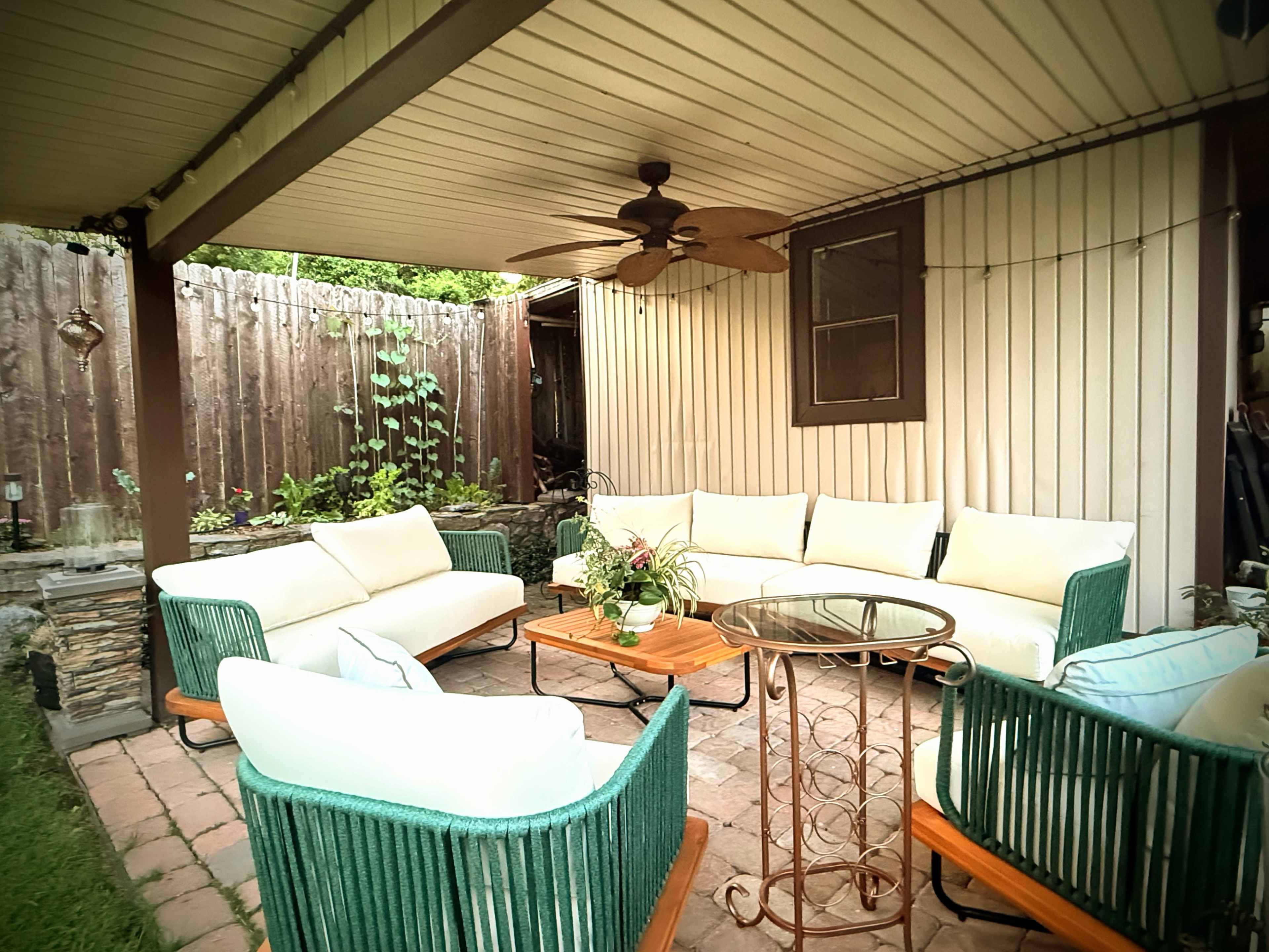 The image shows a covered outdoor patio area furnished with white cushioned seating and a glass-top coffee table surrounded by green wicker chairs.