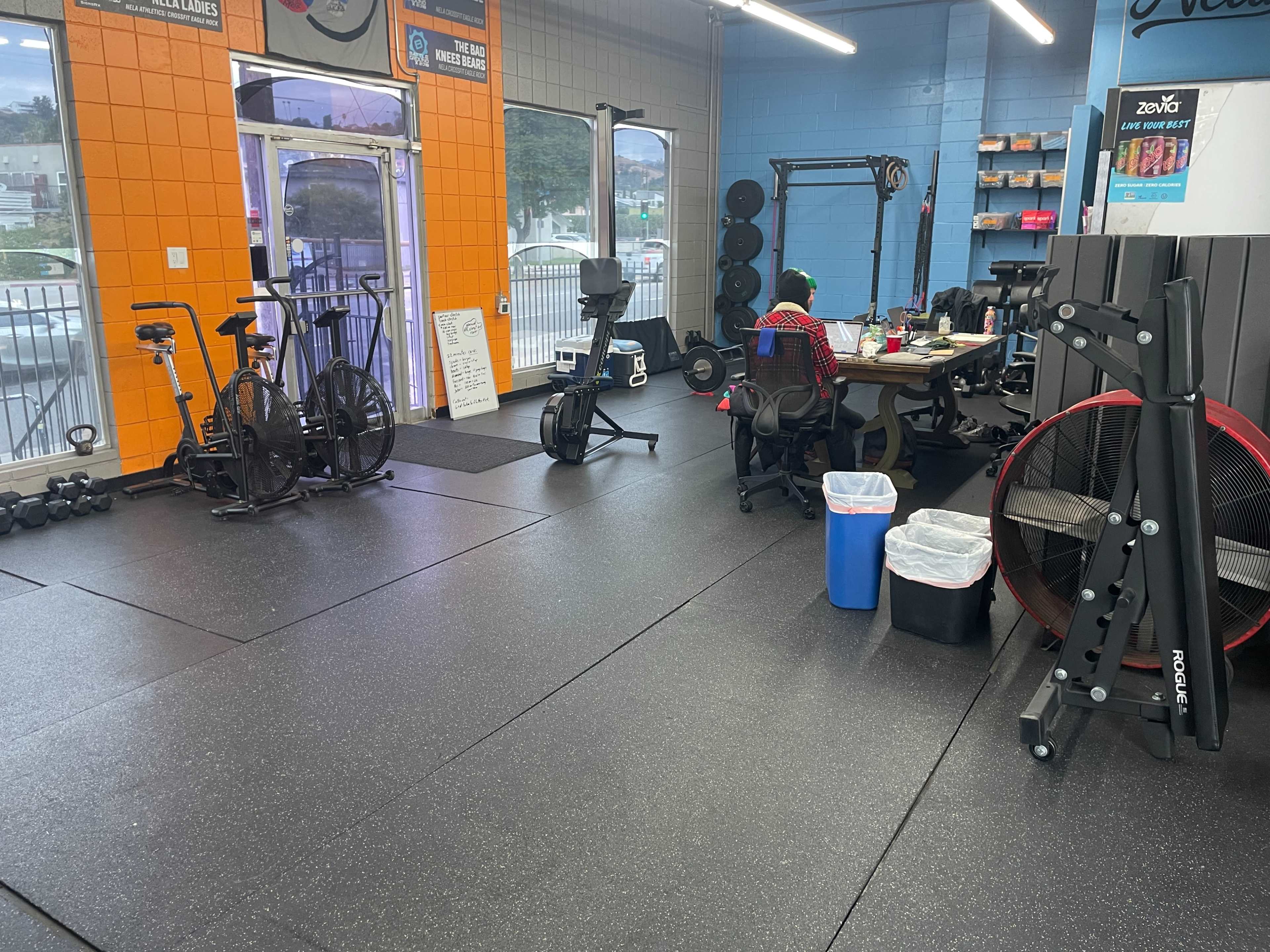 A gym interior featuring exercise equipment, including stationary bikes and weights, with a workstation and garbage bin in the corner.