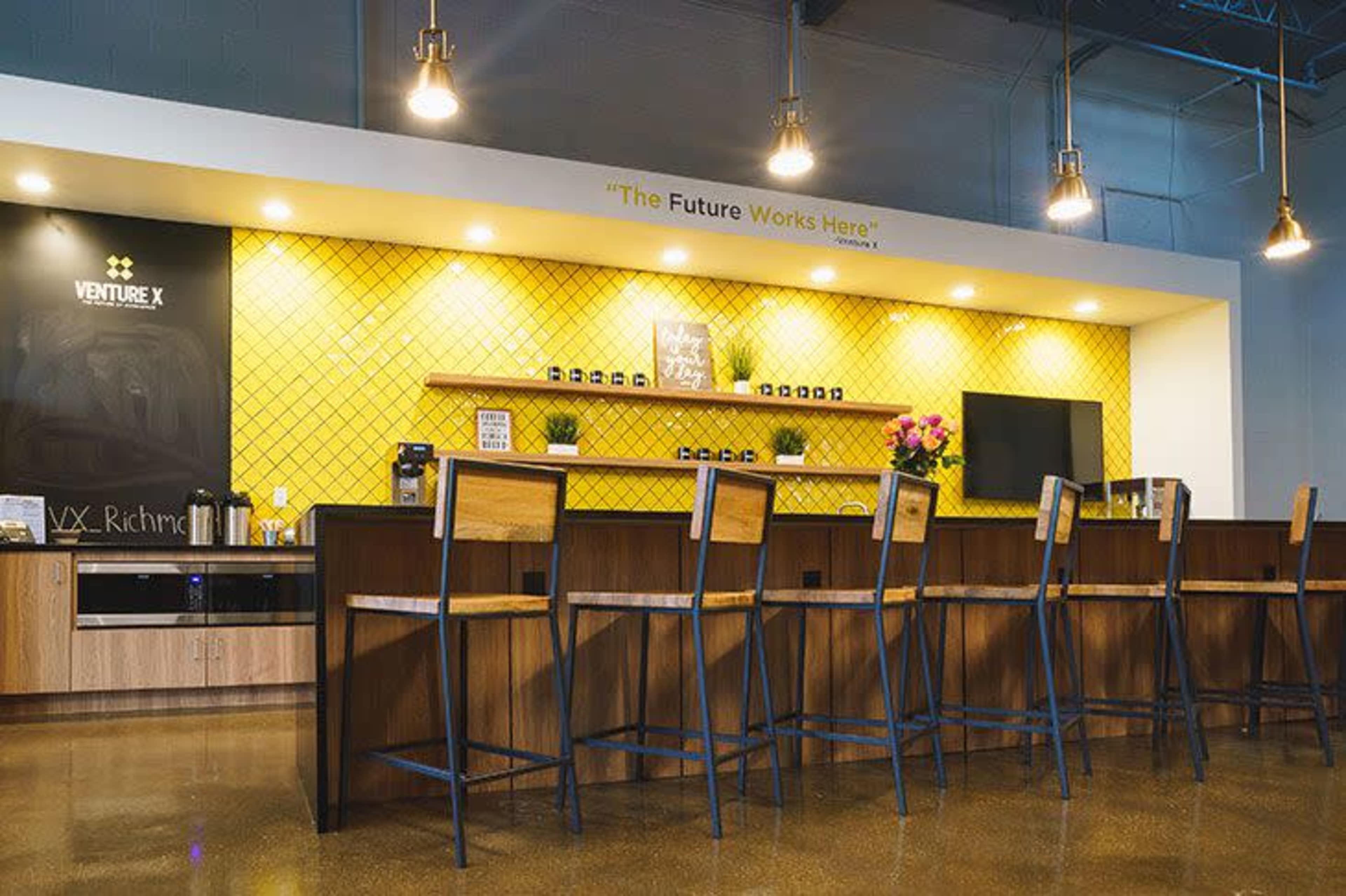 A modern coworking space features a long bar with high stools, yellow tiled wall accents, and a television mounted above a shelf with plants and coffee supplies.