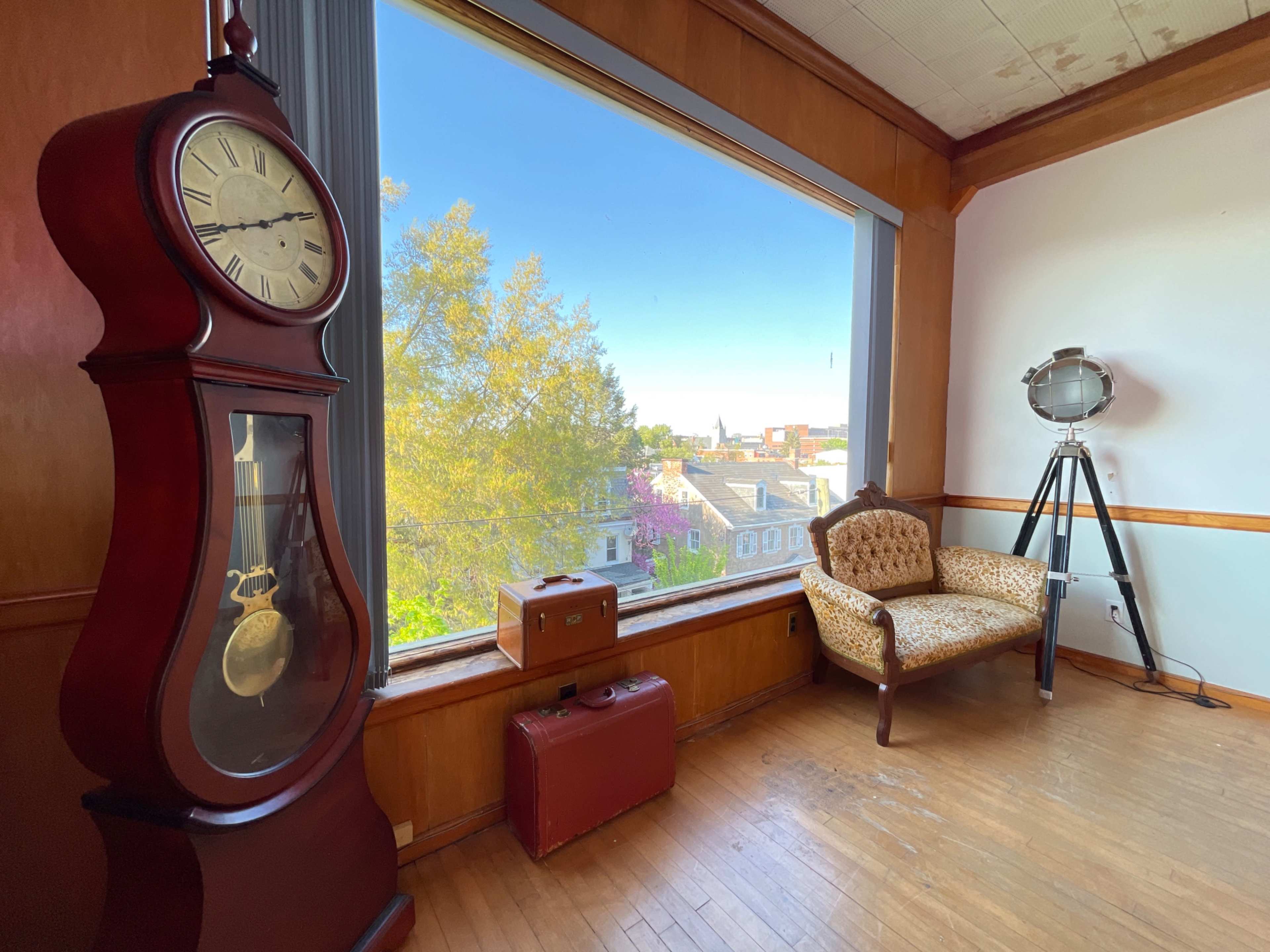 A room with a large window overlooking a cityscape, featuring a tall grandfather clock, a vintage suitcase, and a chair beside a tripod spotlight.