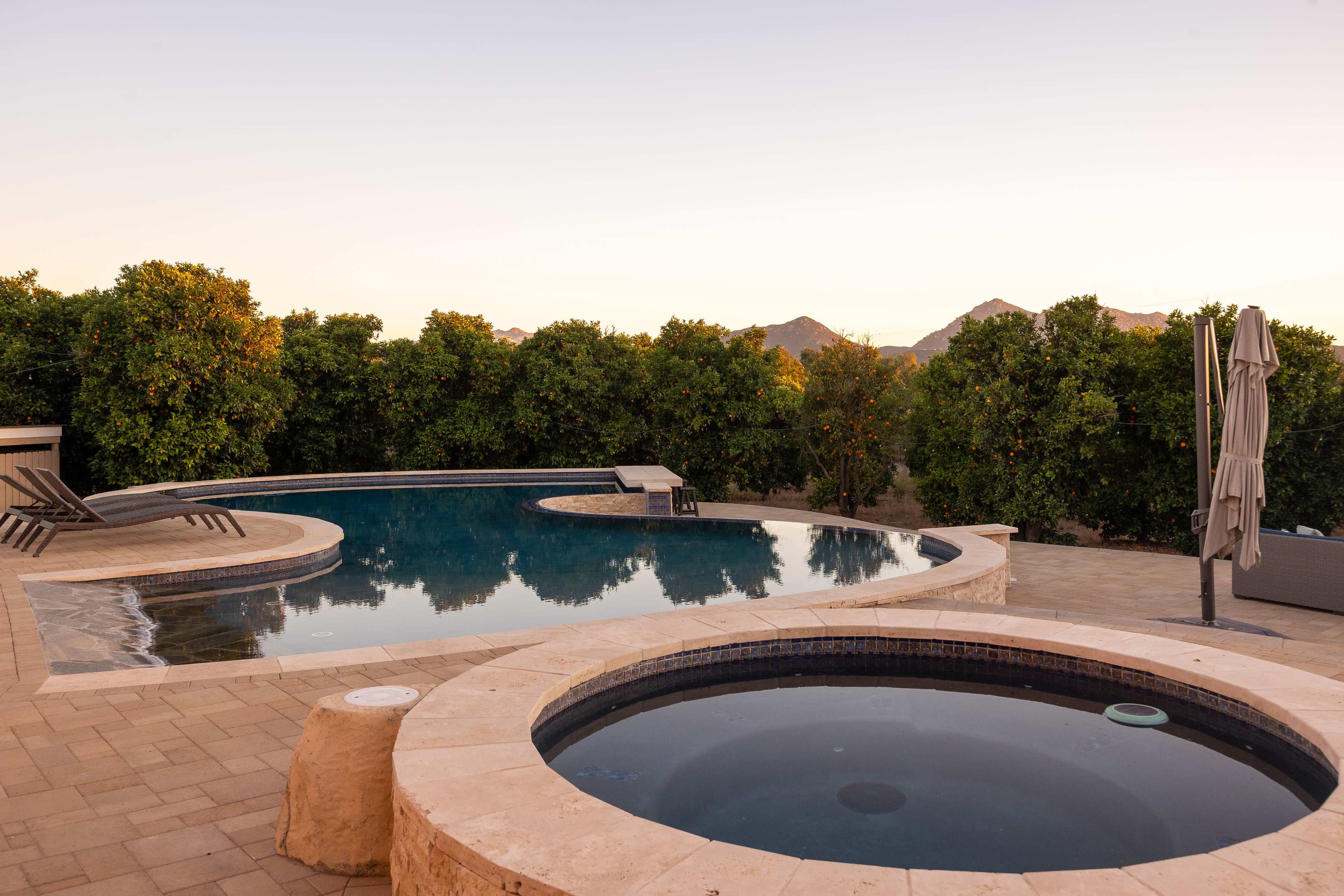 The image shows a modern residential backyard featuring a large swimming pool and a separate hot tub surrounded by greenery and mountains in the background.