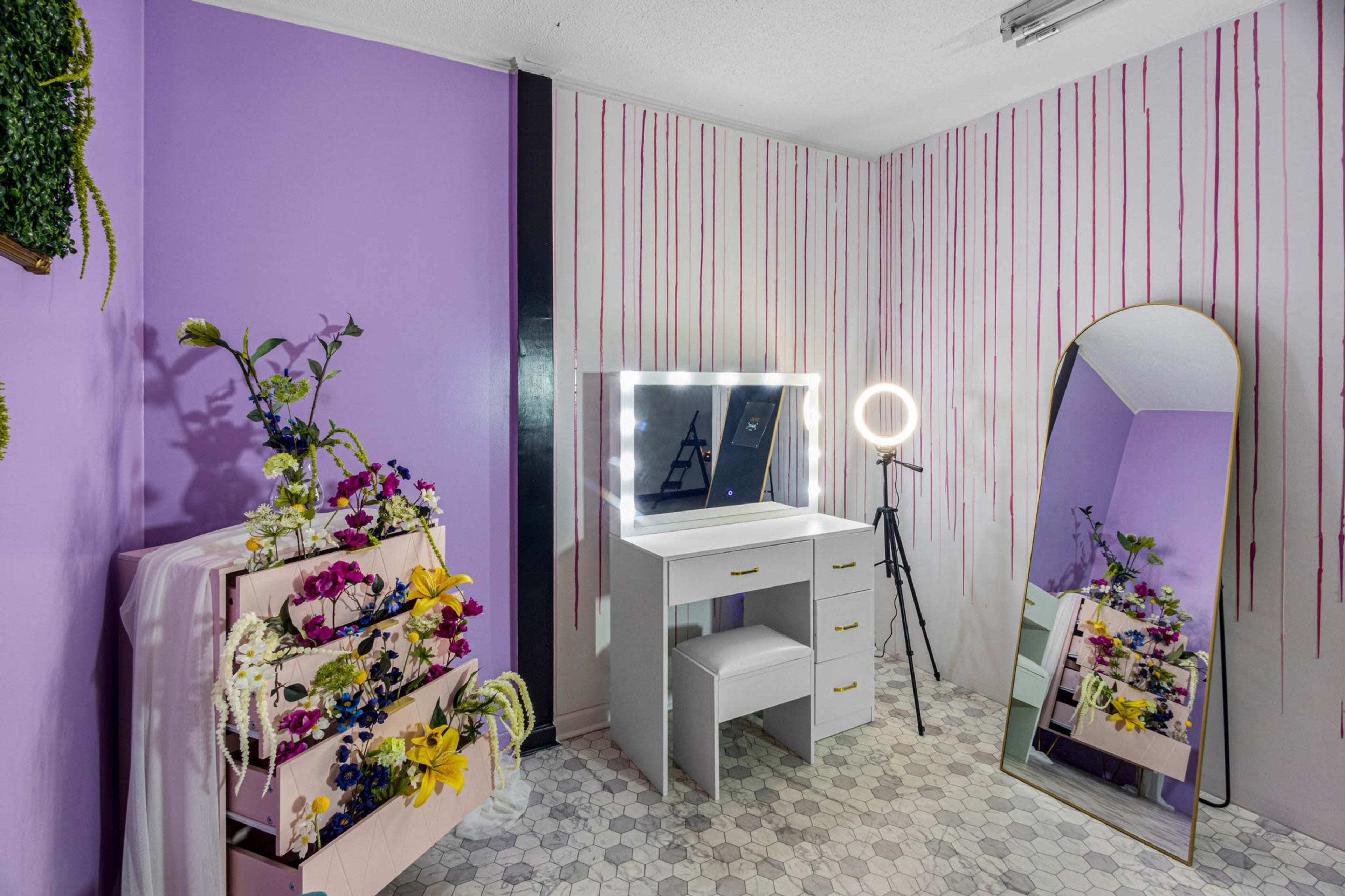 A modern vanity area with a mirror, a ring light, and a chest of drawers decorated with flowers against a purple wall.