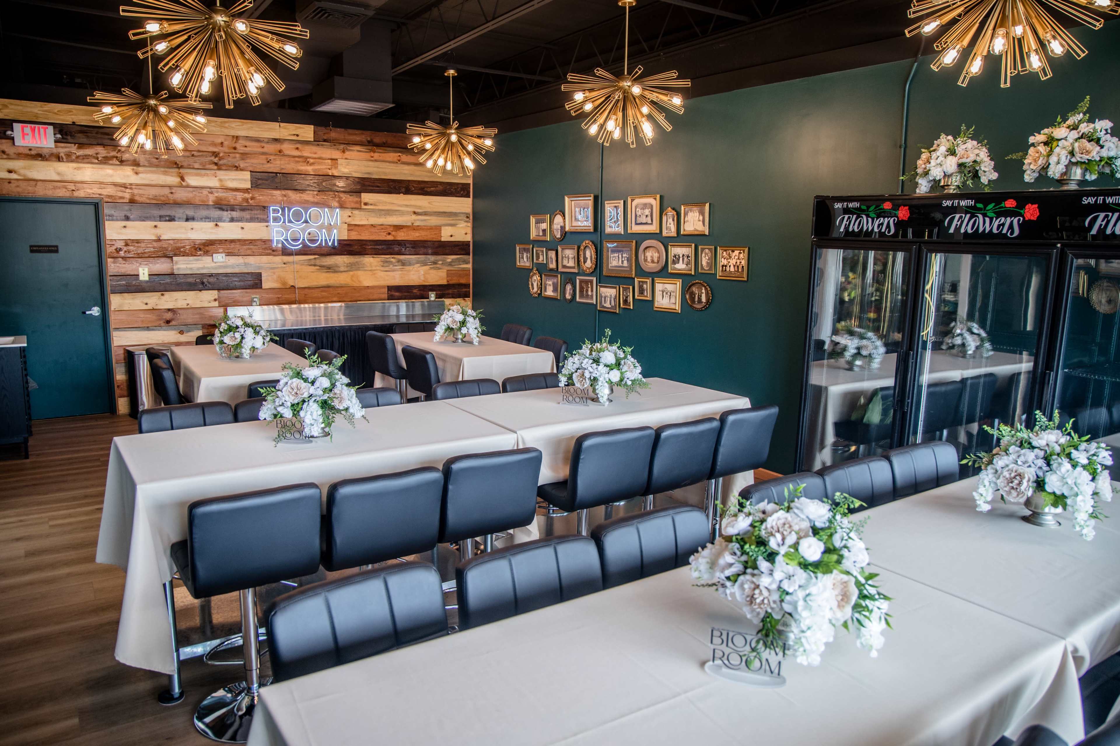 The image shows a stylish interior of a dining space featuring tables with white tablecloths and floral centerpieces, surrounded by wooden accents on the walls and decorative lighting fixtures.