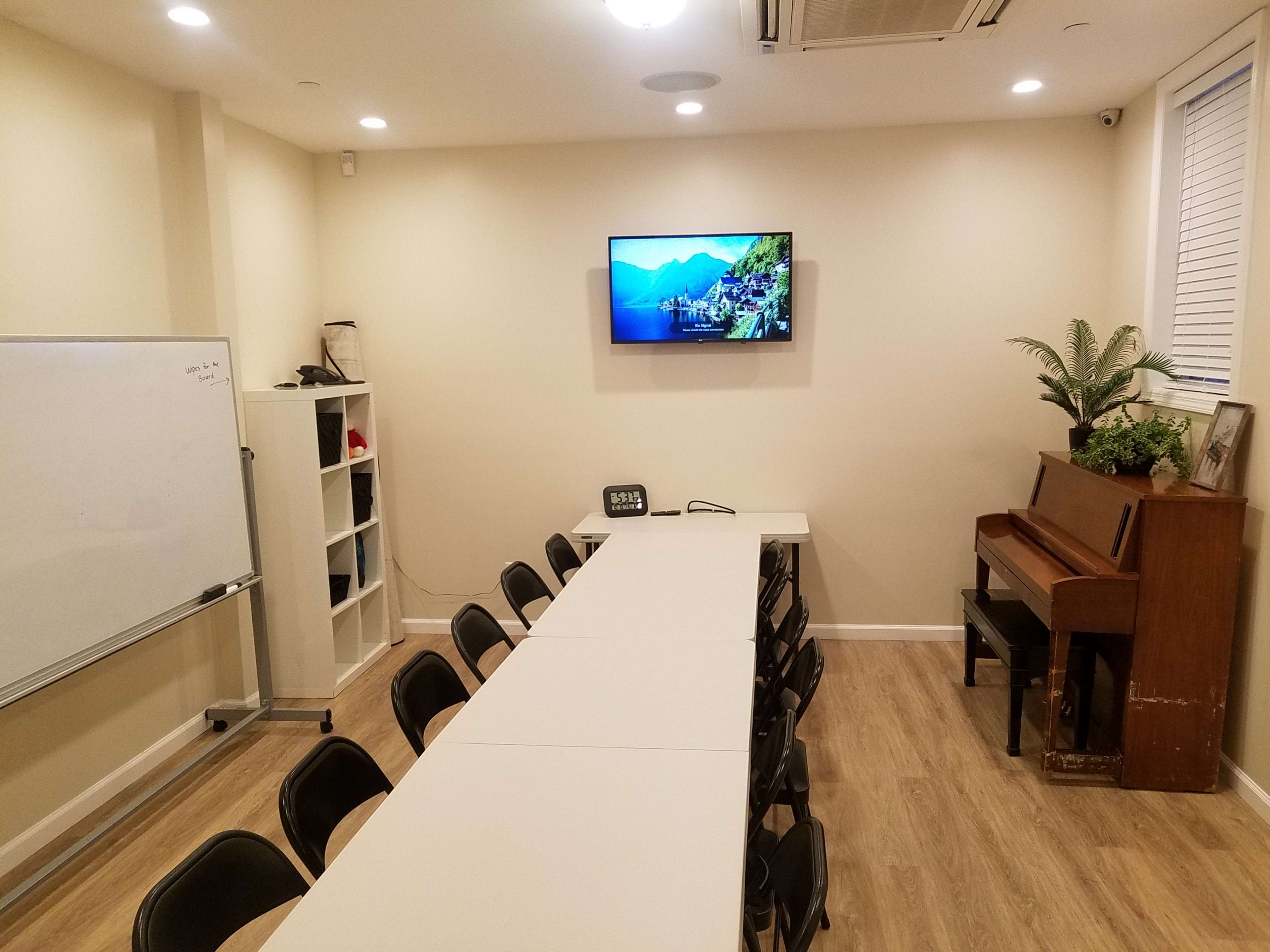 A meeting room features a long table surrounded by chairs, a wall-mounted TV displaying a landscape, a whiteboard, a bookshelf, and a piano in one corner.