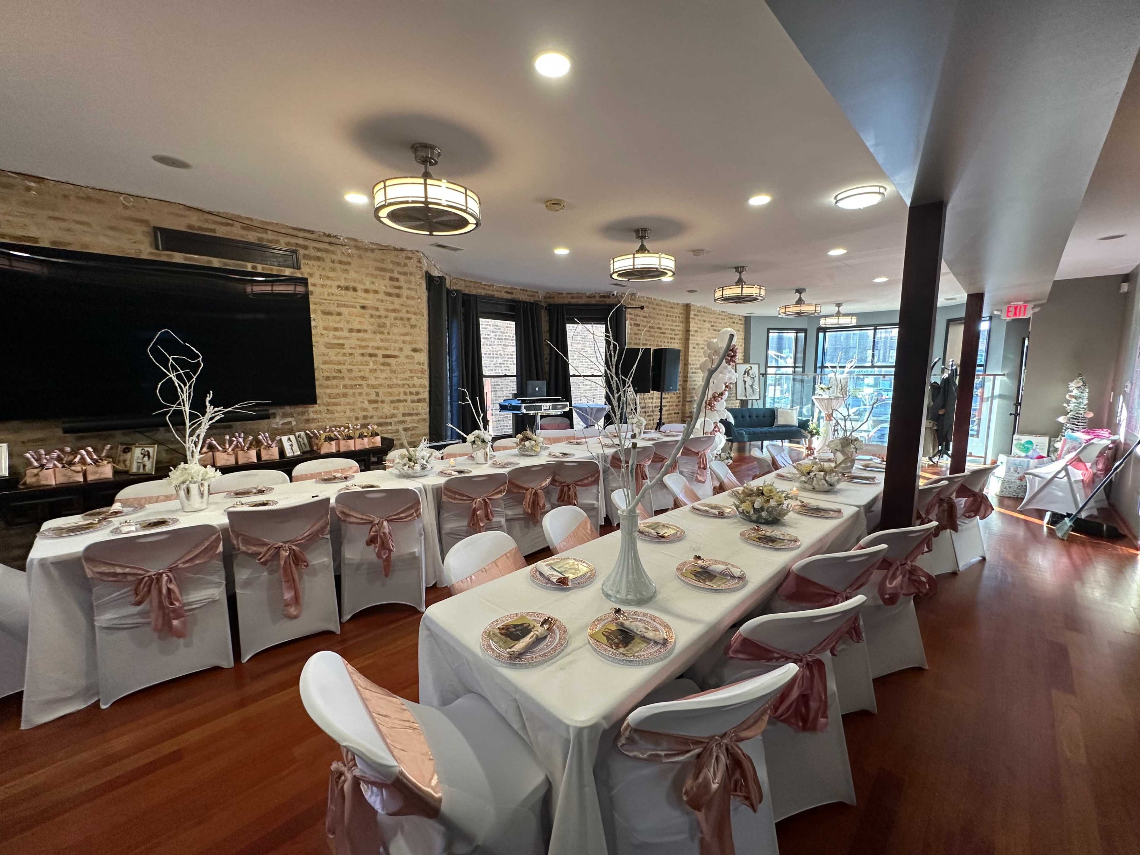 A dining area set for an event, featuring long tables adorned with white tablecloths, pink satin chair sashes, and decorative centerpieces.