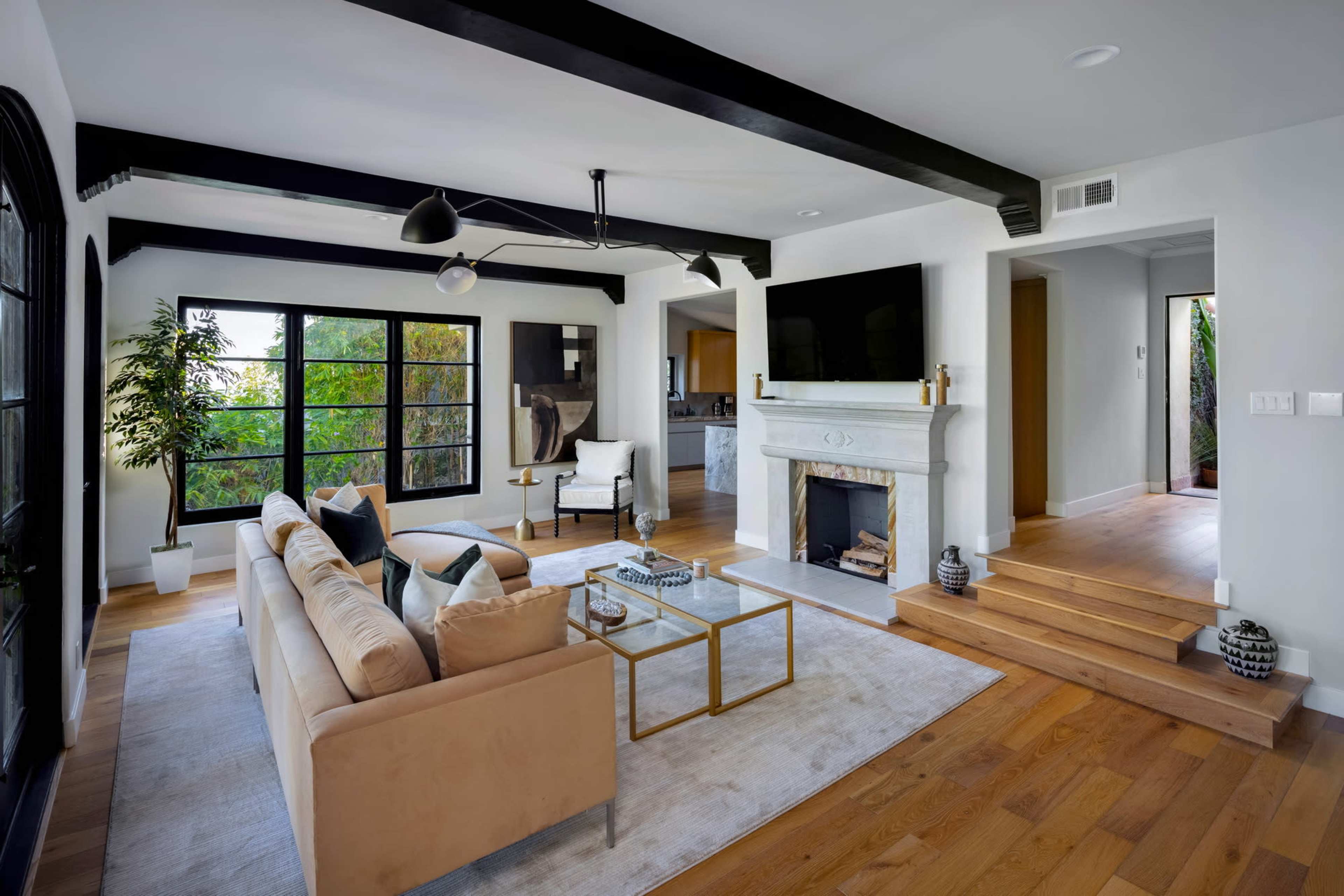 A modern living room with a beige sofa, a glass coffee table, a fireplace, and large windows overlooking greenery.