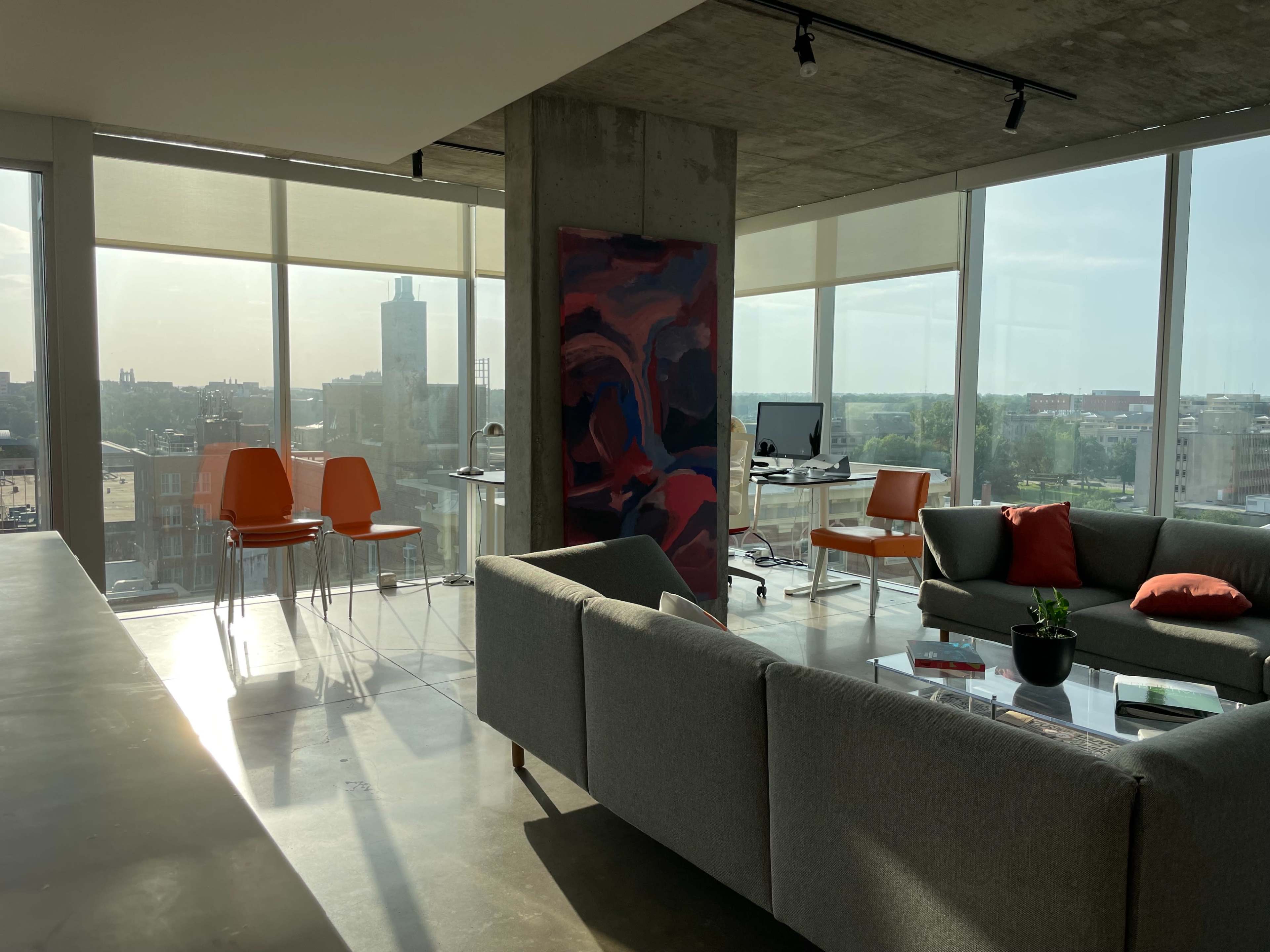 An open office space features a gray couch, orange chairs, a desk with a computer, and large windows that offer a view of the city.