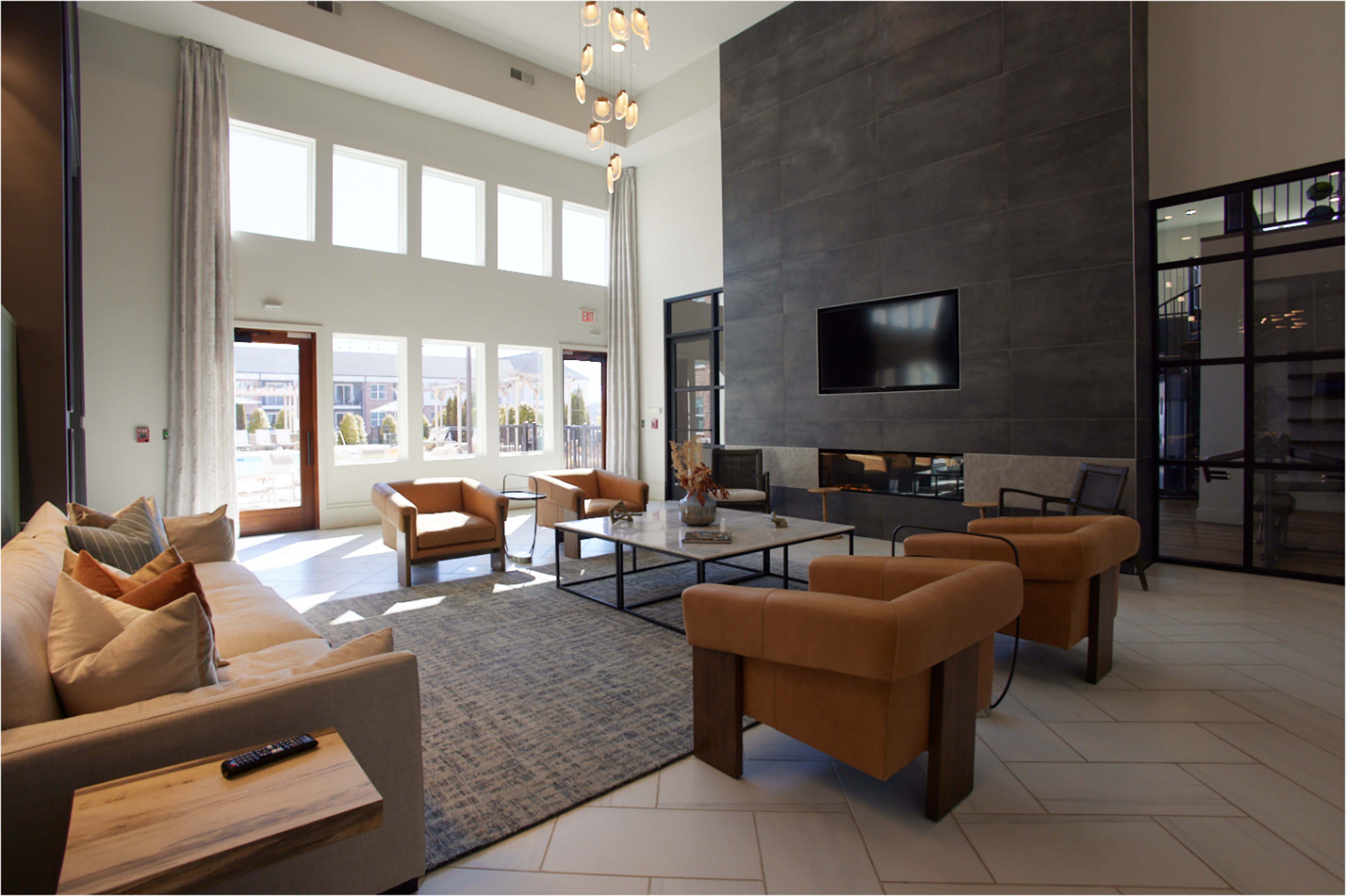 The image depicts a modern lounge area with a high ceiling, featuring a large stone wall, a television mounted above a contemporary fireplace, and seating arranged around a coffee table.