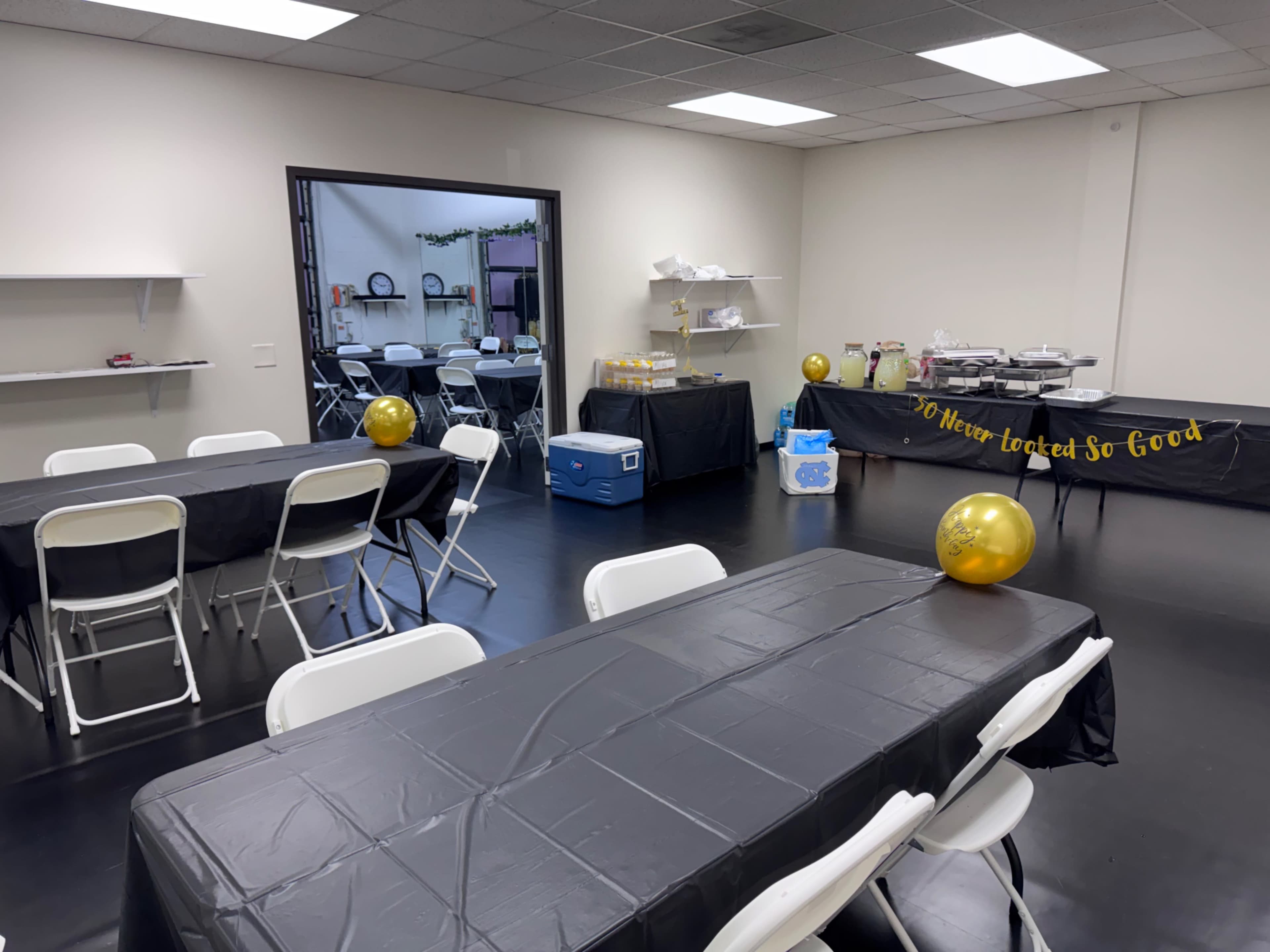 The image shows a decorated event space with black tablecloths spread across long tables, some balloons, and a buffet setup against one wall.