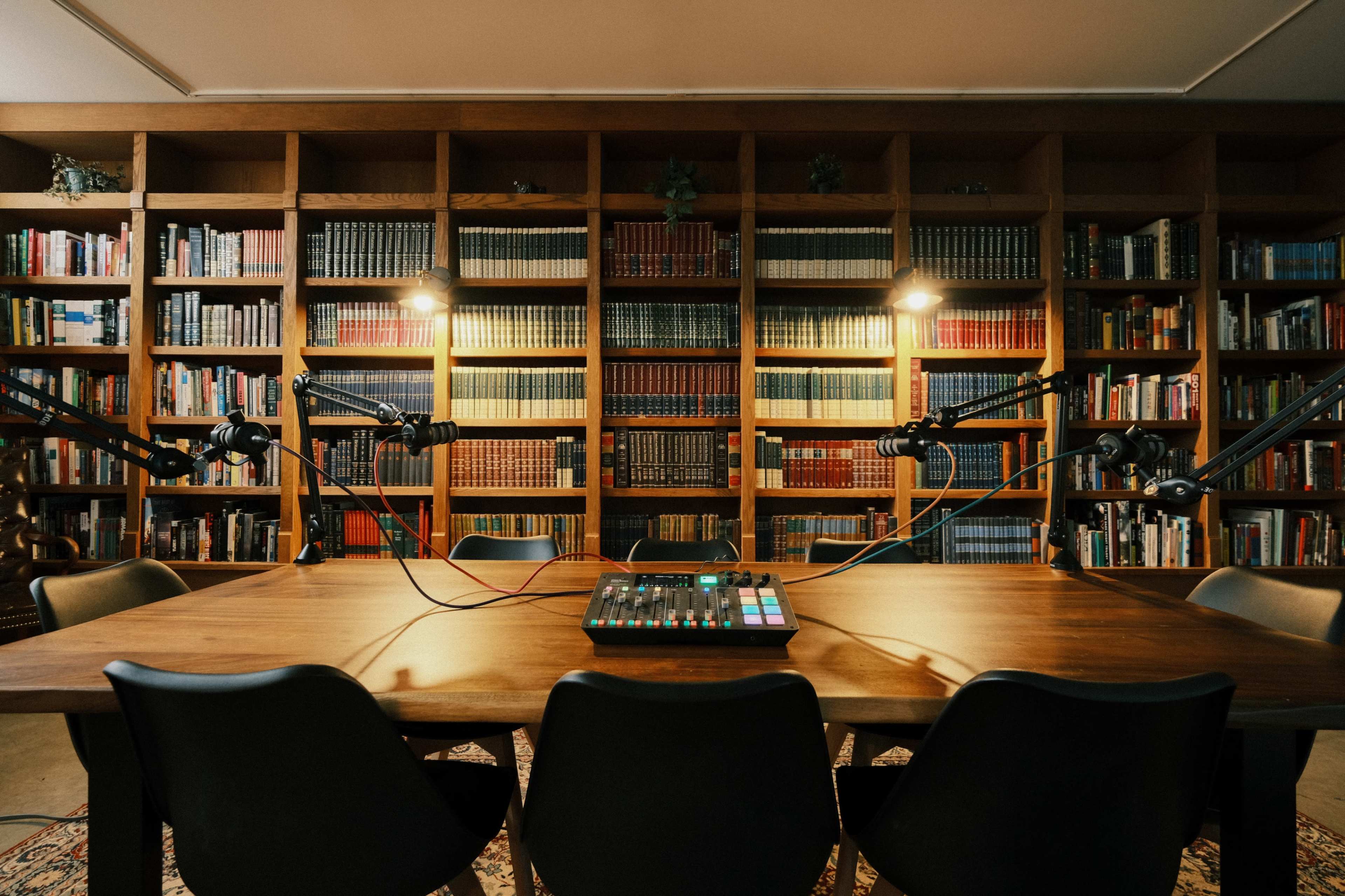 A well-lit podcasting studio features a wooden table with audio equipment and microphones, surrounded by shelves filled with books.
