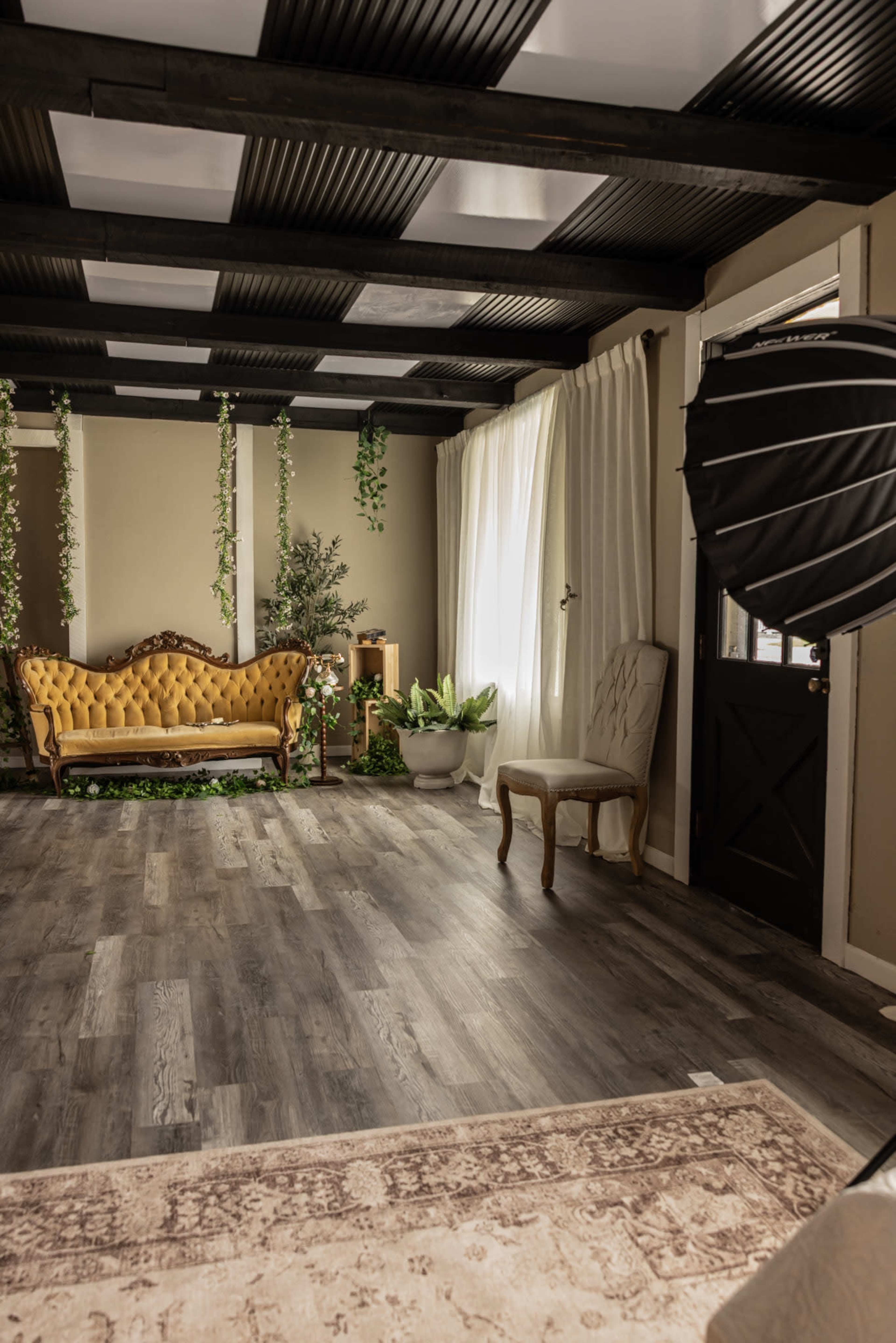 The image shows a stylish interior room featuring a vintage yellow sofa, potted plants, a wooden chair, and a photography lighting setup.