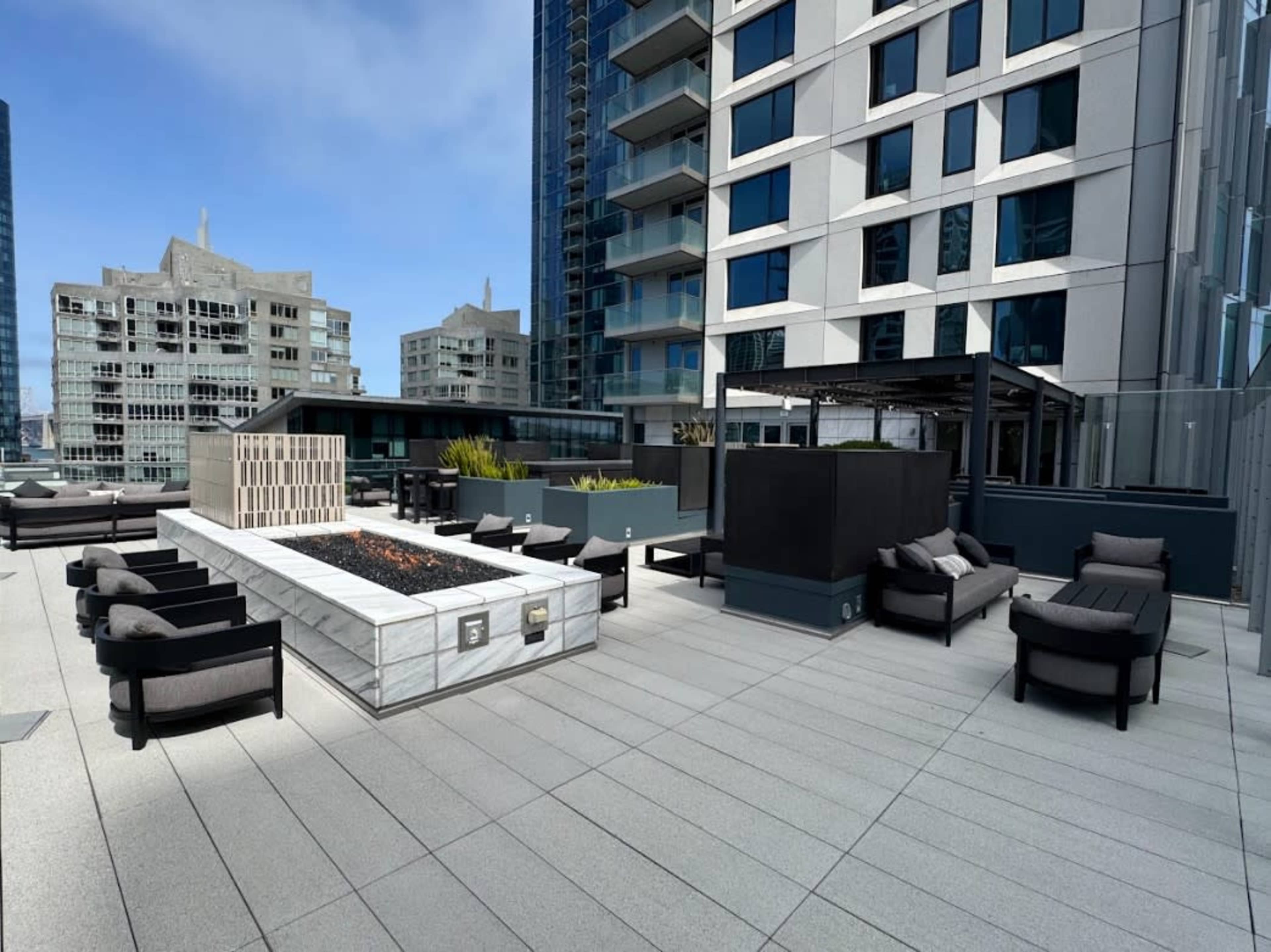 The image shows a modern rooftop terrace featuring seating areas, a fire pit, and surrounding high-rise buildings.