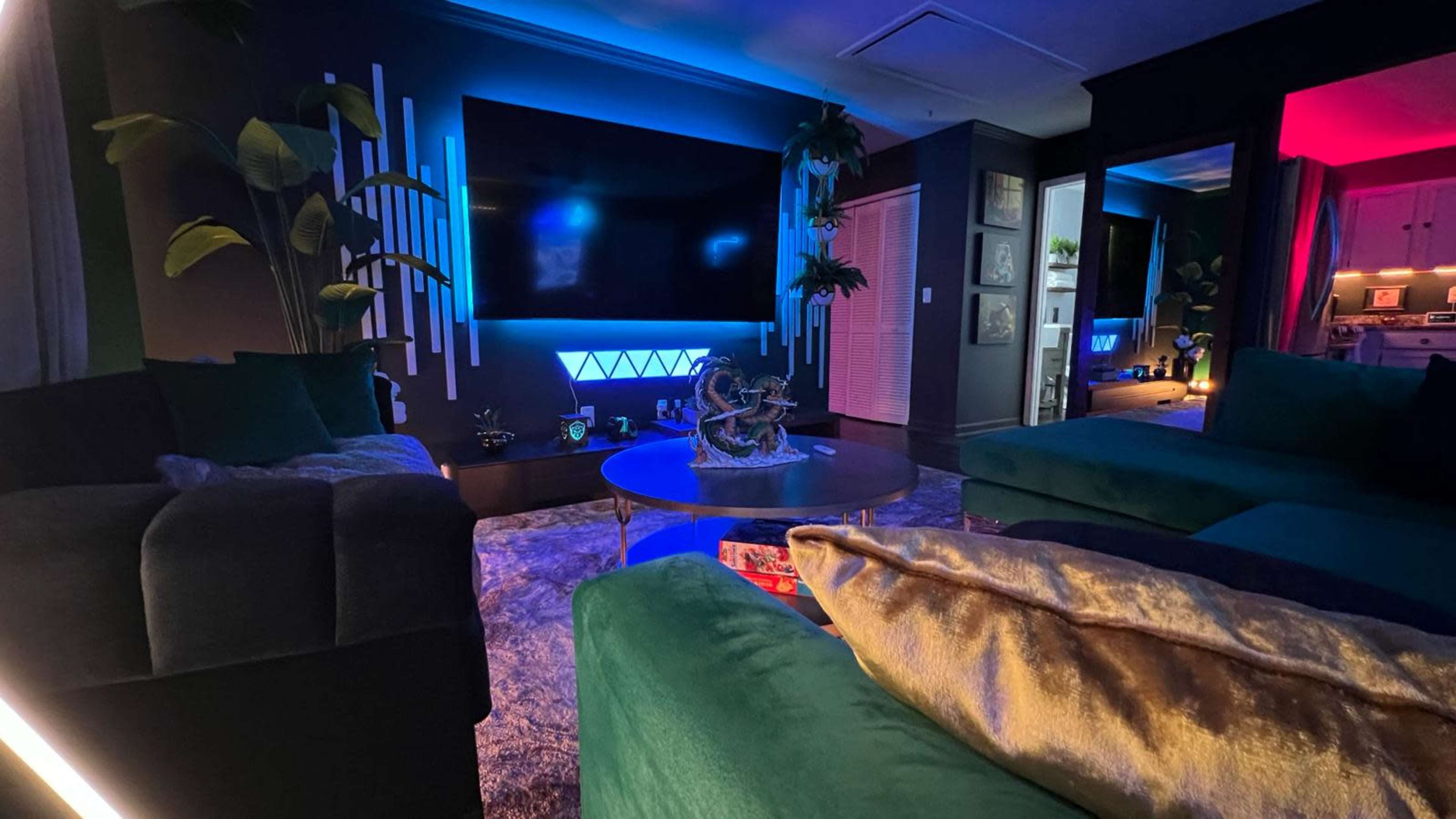 The room features a modern design with green and blue lighting, a large wall-mounted TV, and plush seating arranged around a decorative coffee table.