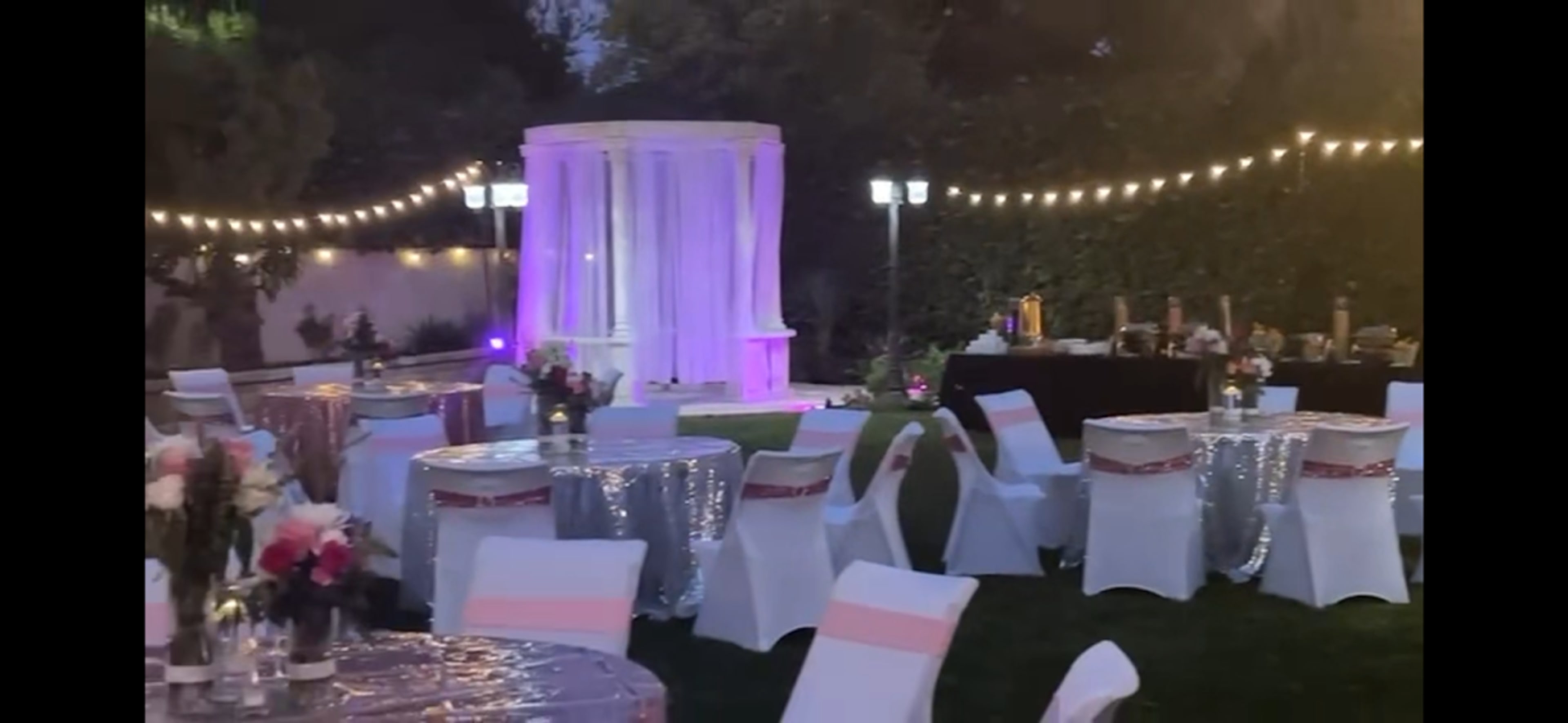 The image shows a beautifully arranged outdoor event space with decorated tables, white chairs, and a gazebo illuminated by purple lights, surrounded by string lights.