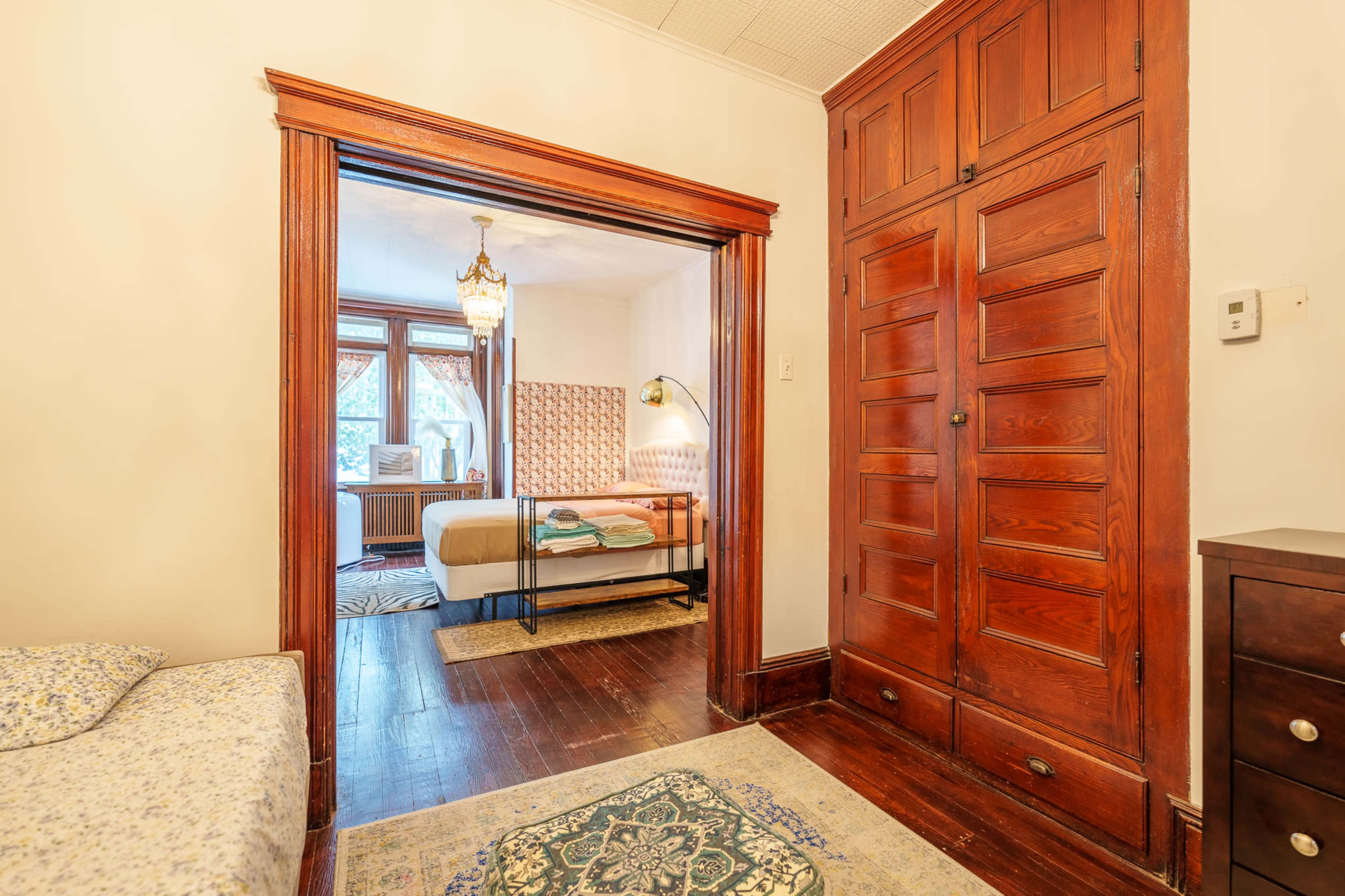 The image shows a hallway leading into a bedroom with a wooden door frame, featuring a bed in the background, an ornate light fixture, and wooden cabinetry.