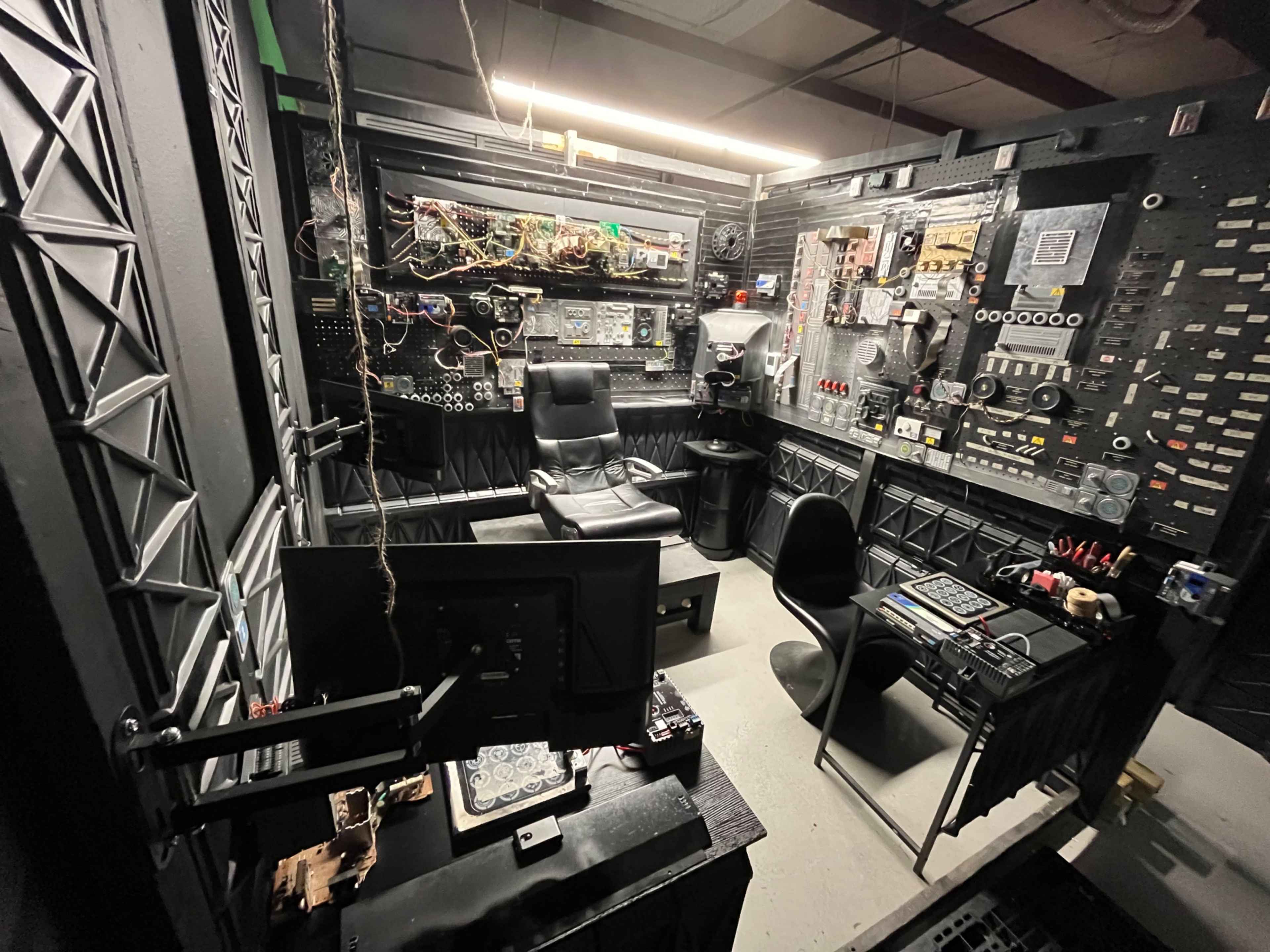 The image shows a dimly lit control room filled with various electronic panels, wires, and equipment, featuring multiple chairs and a monitor setup.