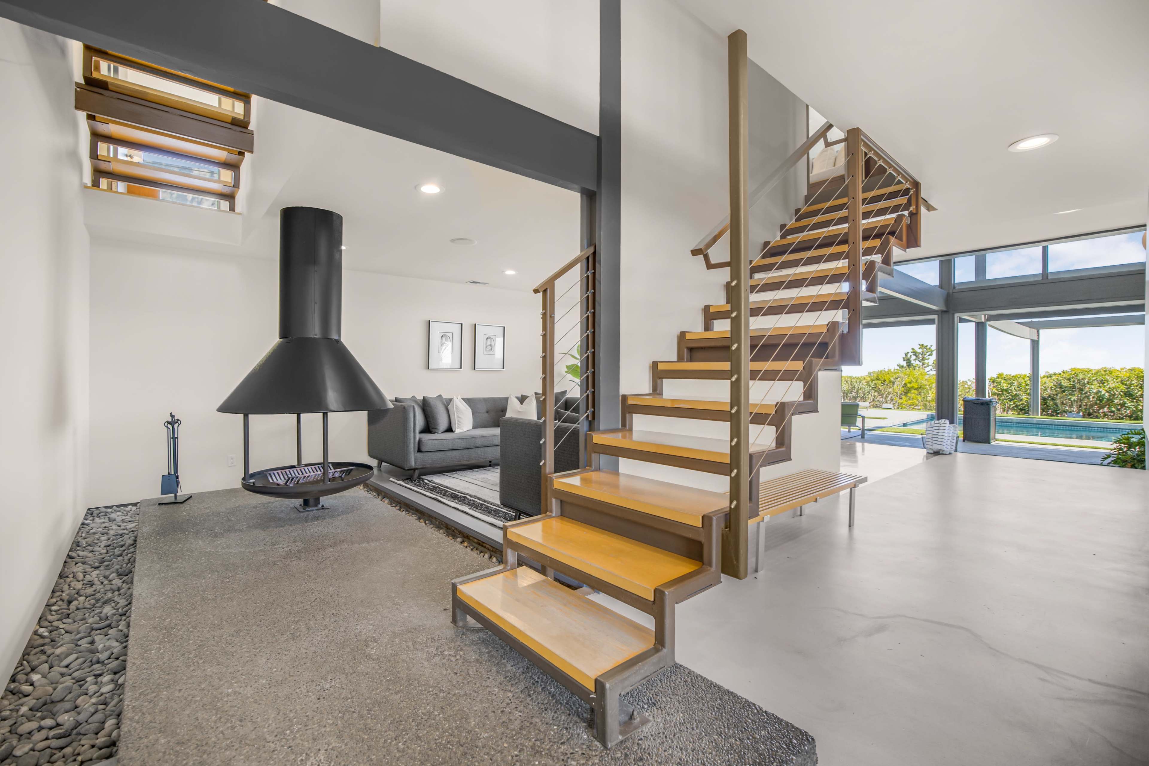 The image shows a modern interior featuring a staircase leading to an upper level, a circular fireplace, and large windows allowing natural light to illuminate the space.