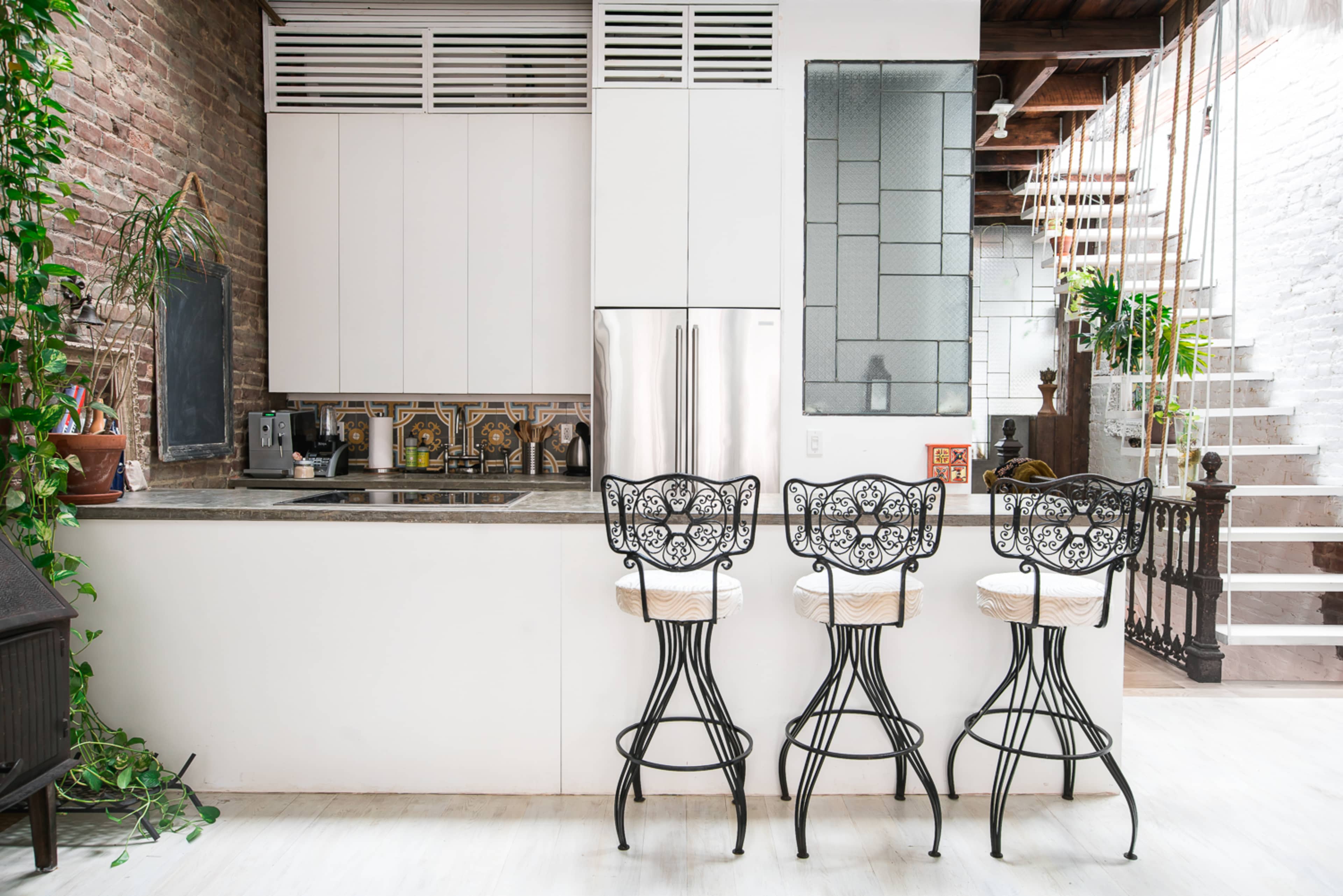 A modern kitchen features a white counter and cabinets with a patterned backsplash, complemented by three wrought-iron bar stools.