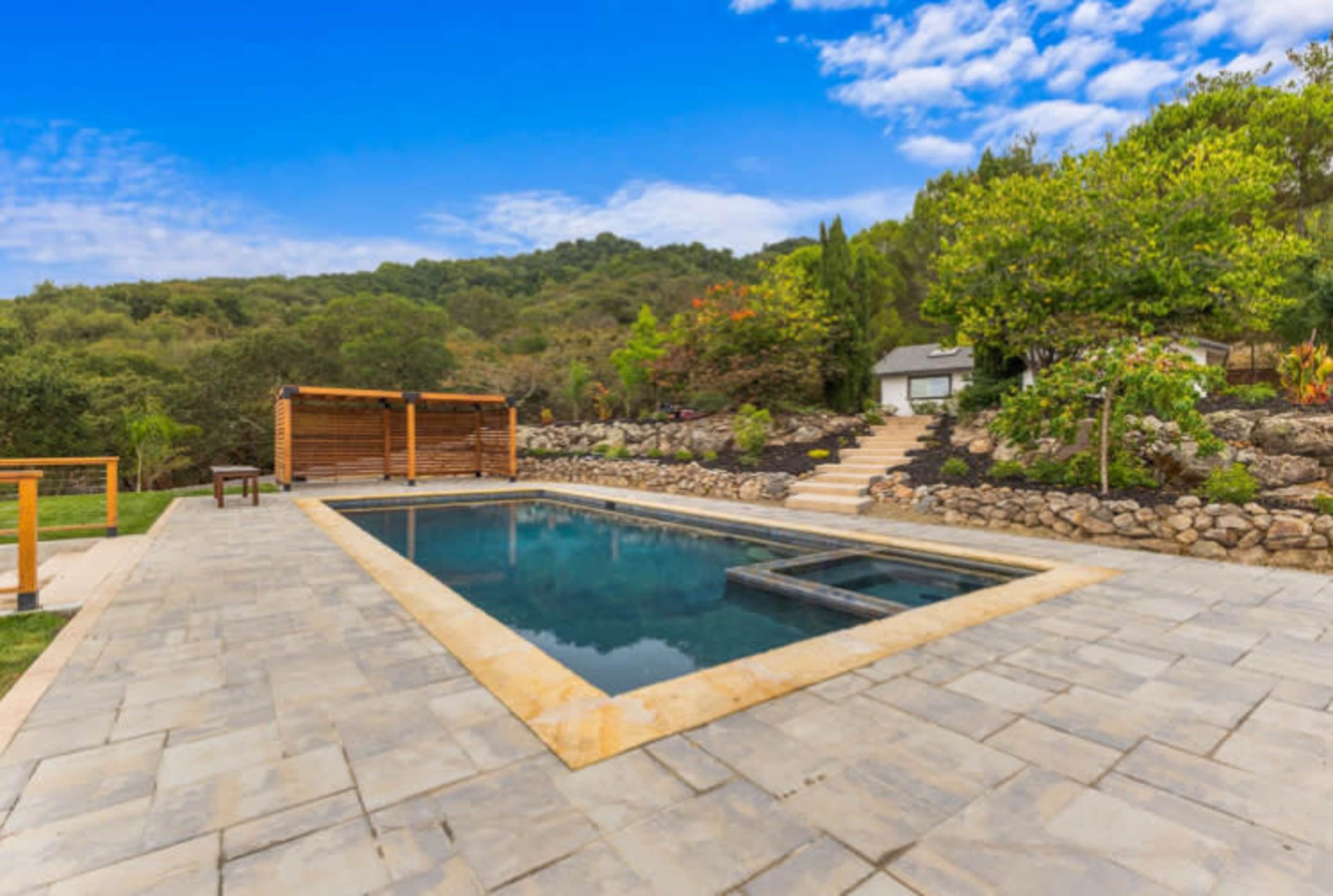A rectangular swimming pool is surrounded by stone pavement and landscaped gardens under a partly cloudy sky.