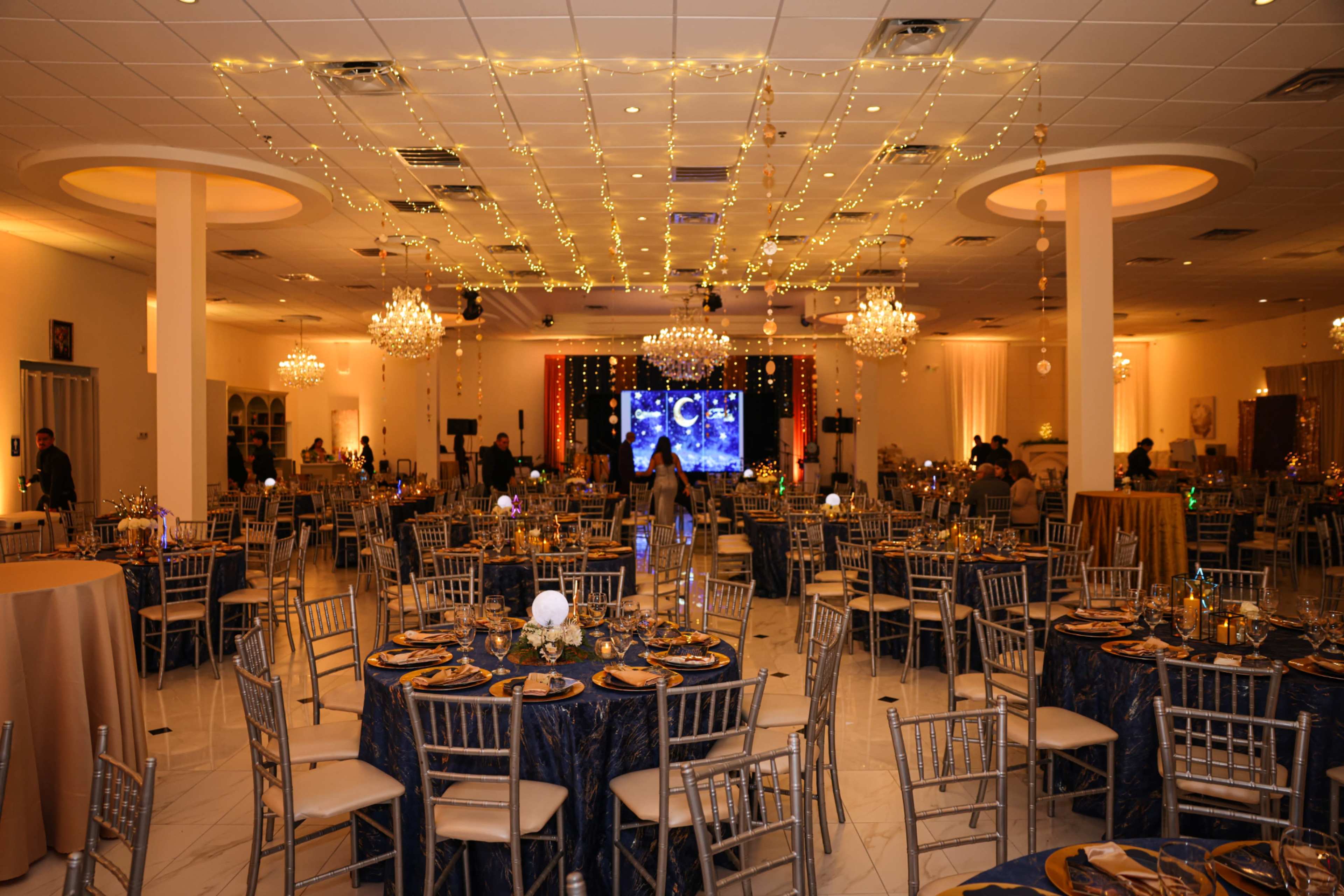 The image shows a banquet hall decorated for an event, featuring round tables set with blue tablecloths and gold accents, chandeliers hanging from the ceiling, and decorative lights strung overhead.