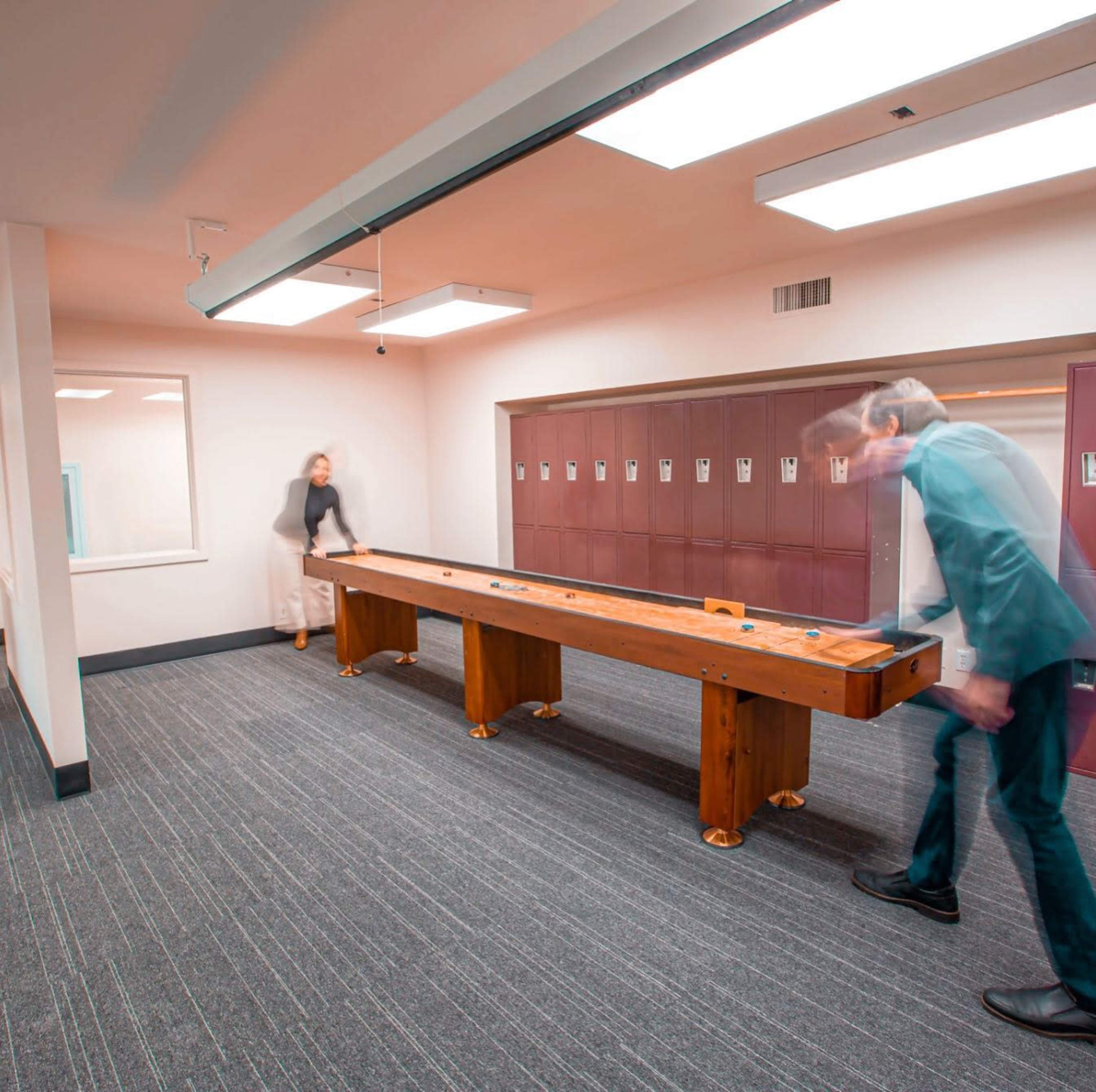 Two people are playing shuffleboard in a room with lockers and overhead lighting.