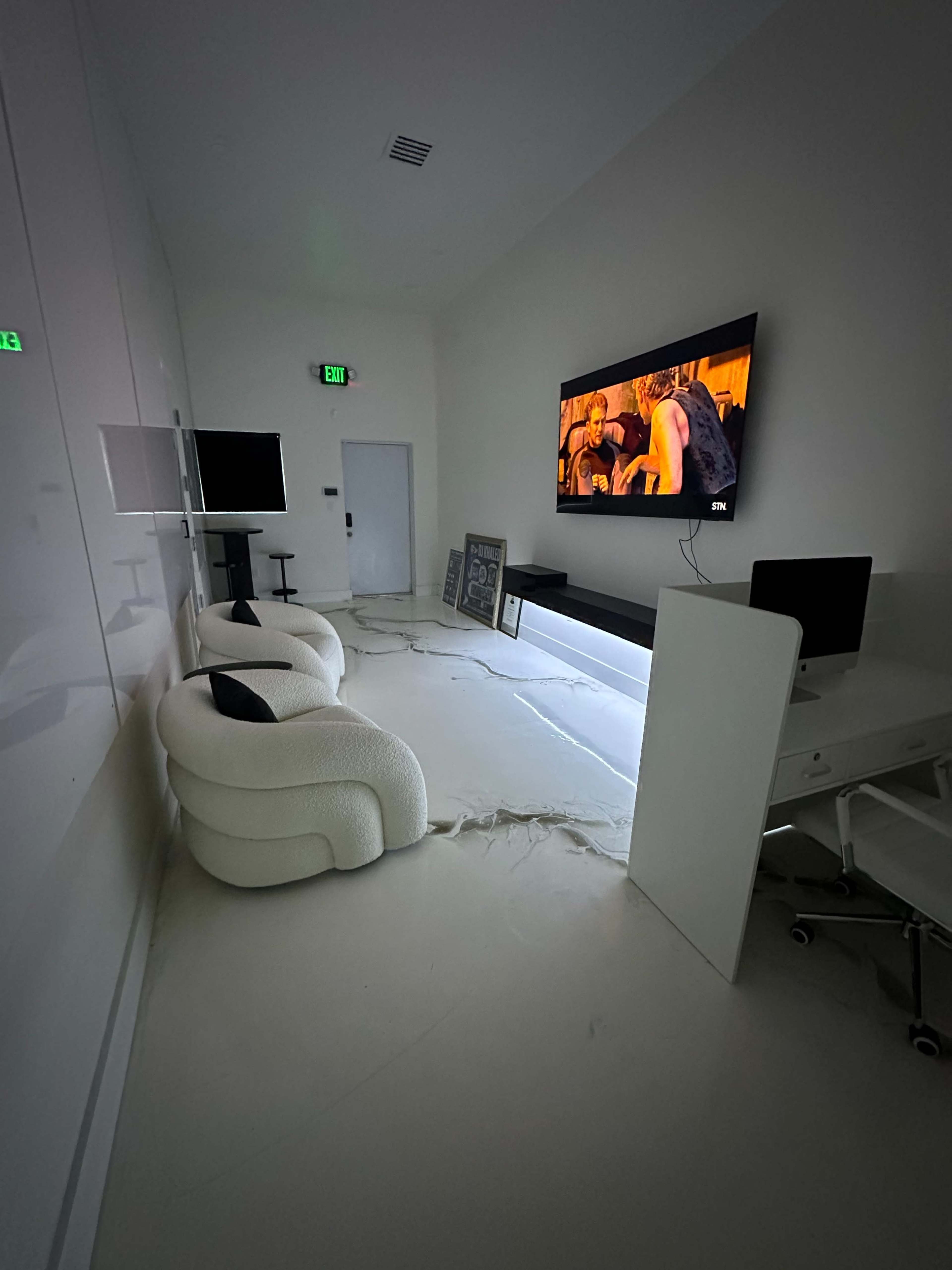 The image shows a modern, minimalistic room featuring two curved white chairs, a wall-mounted television displaying a scene from a movie, and a sleek desk with a computer in front of it.