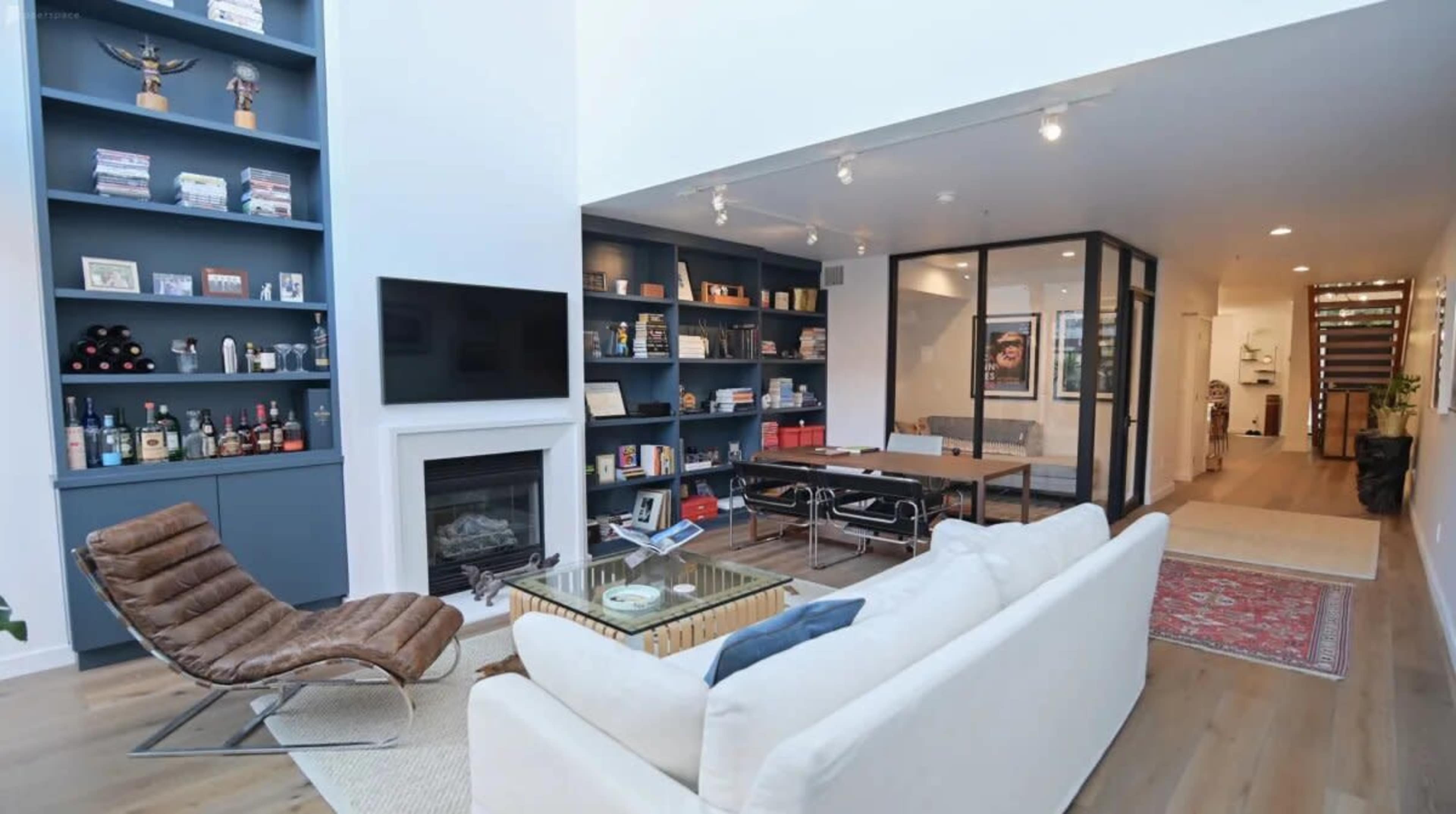 A modern living room with a white sofa, a glass coffee table, a fireplace, a wall-mounted television, and a bookshelf filled with books and decor items, leading into a dining area with a large wooden table.