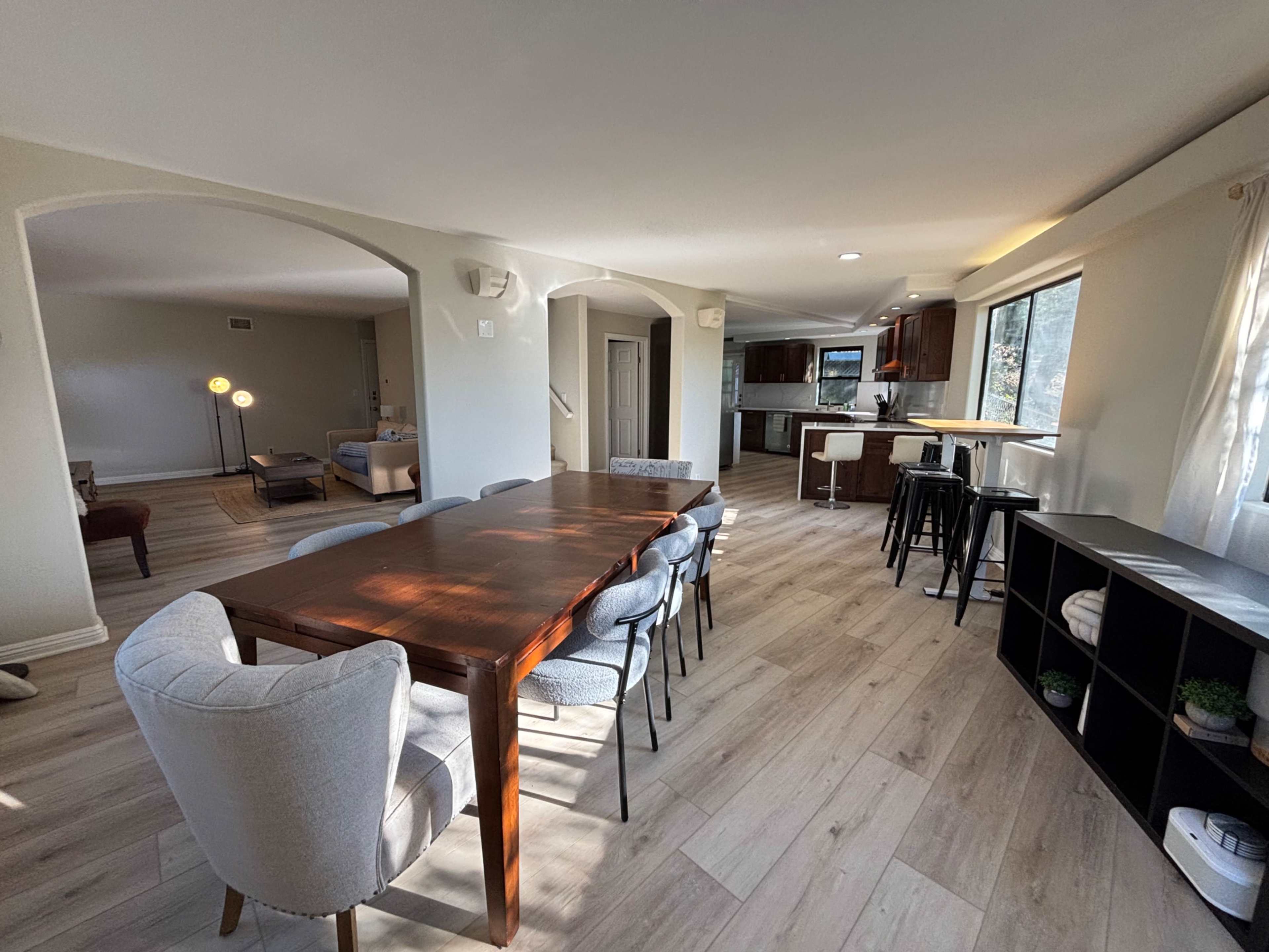 The image shows a spacious dining area with a wooden table and chairs, leading to an open kitchen and living space with large windows.