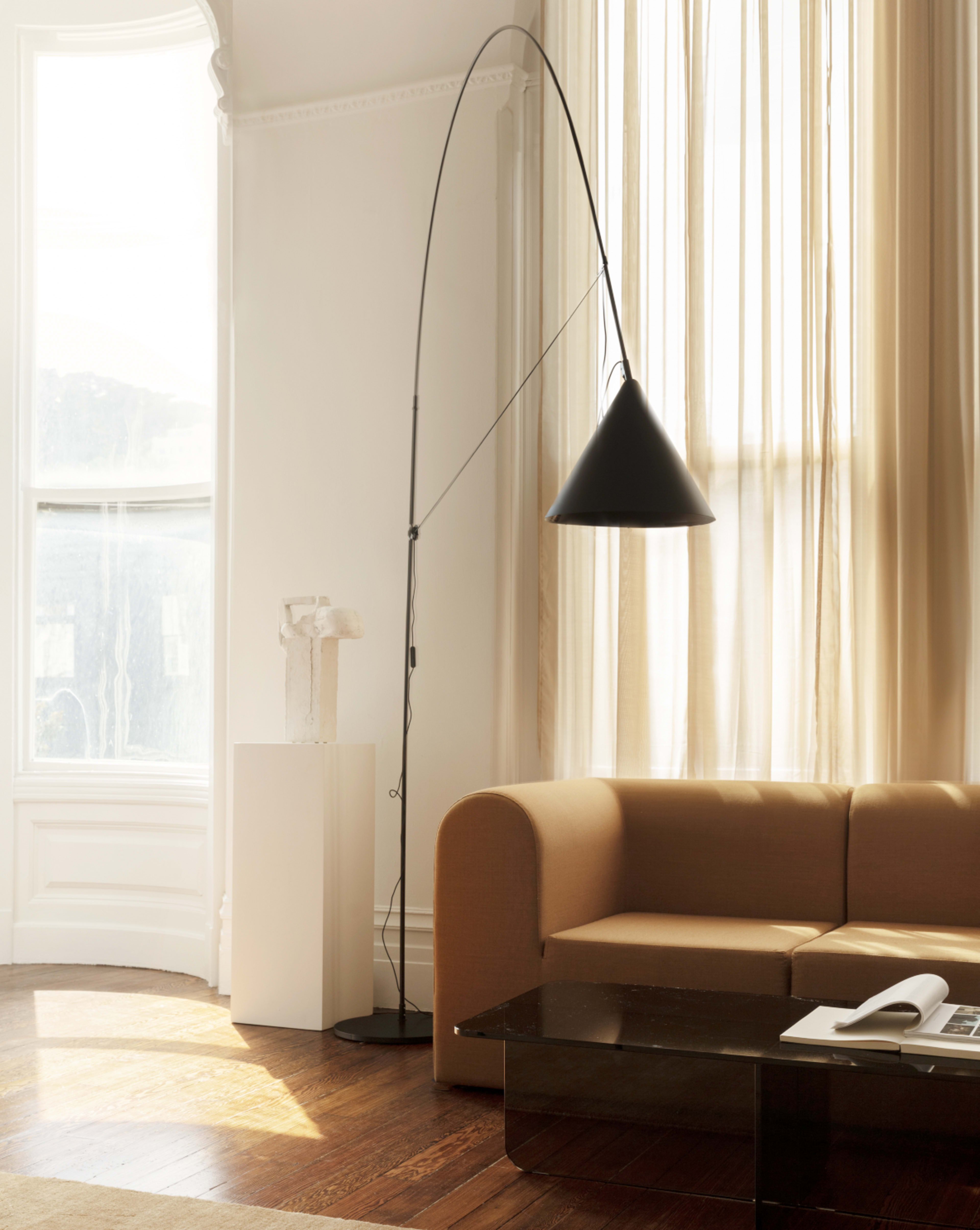 A modern living room features a brown sofa, a curved black floor lamp, a light-colored curtain draped near a large window, and a glass coffee table.
