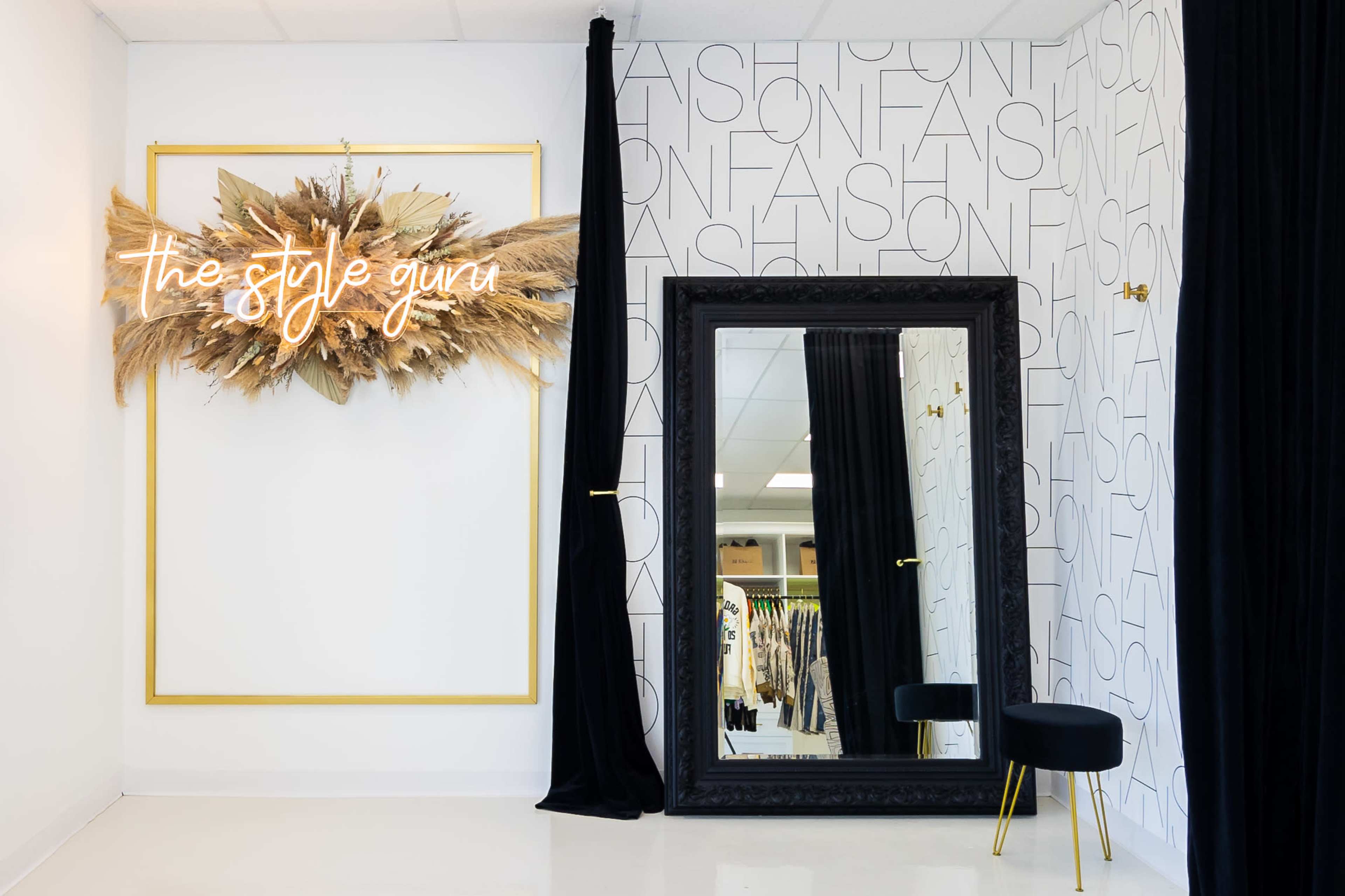 A boutique interior featuring a large mirror, a decorative wall with text, and a wall-mounted sign that reads "the style guru" surrounded by dried floral arrangements.