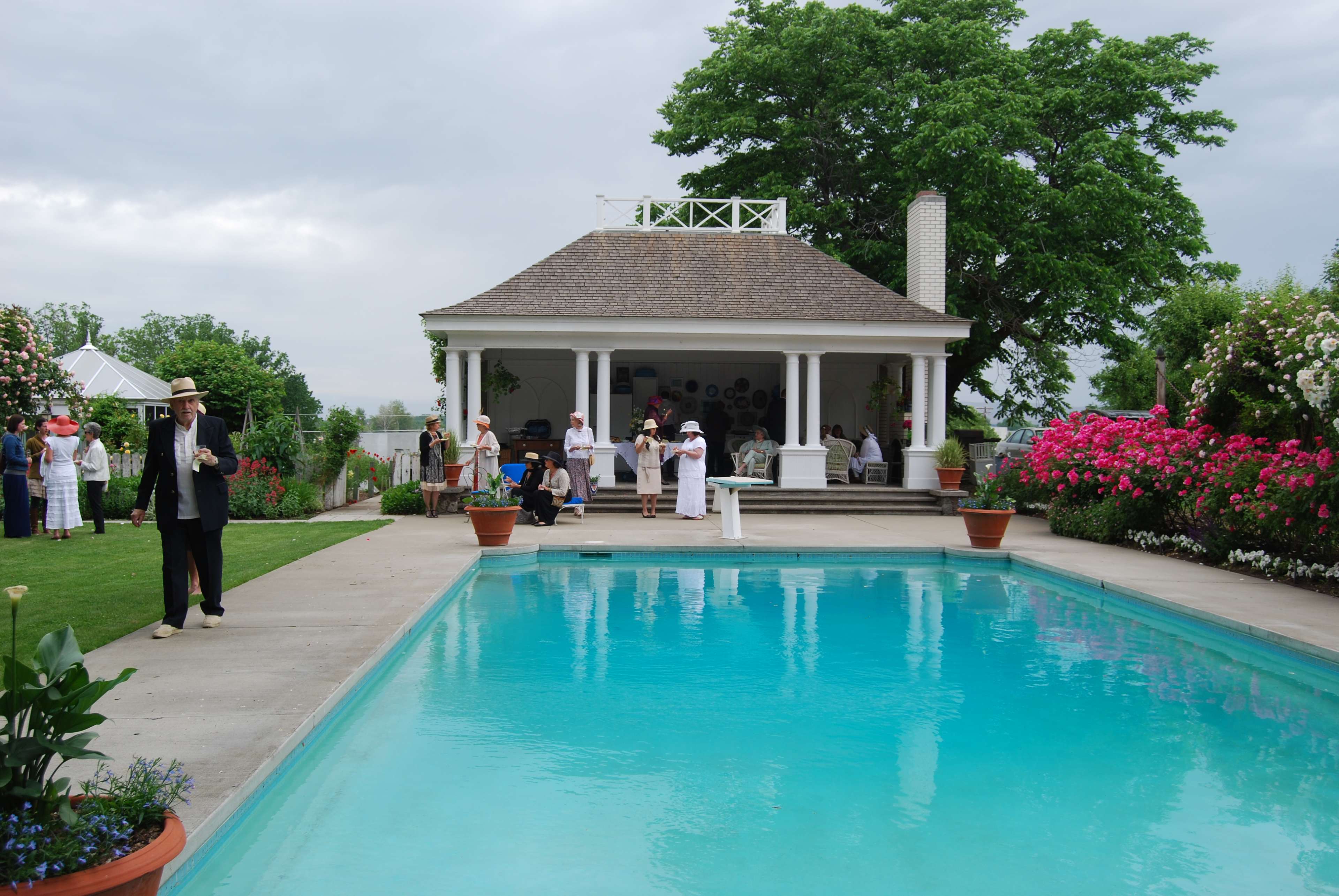 A gathering of people in formal attire attends an outdoor event by a clear swimming pool, surrounded by well-maintained gardens and a gazebo.