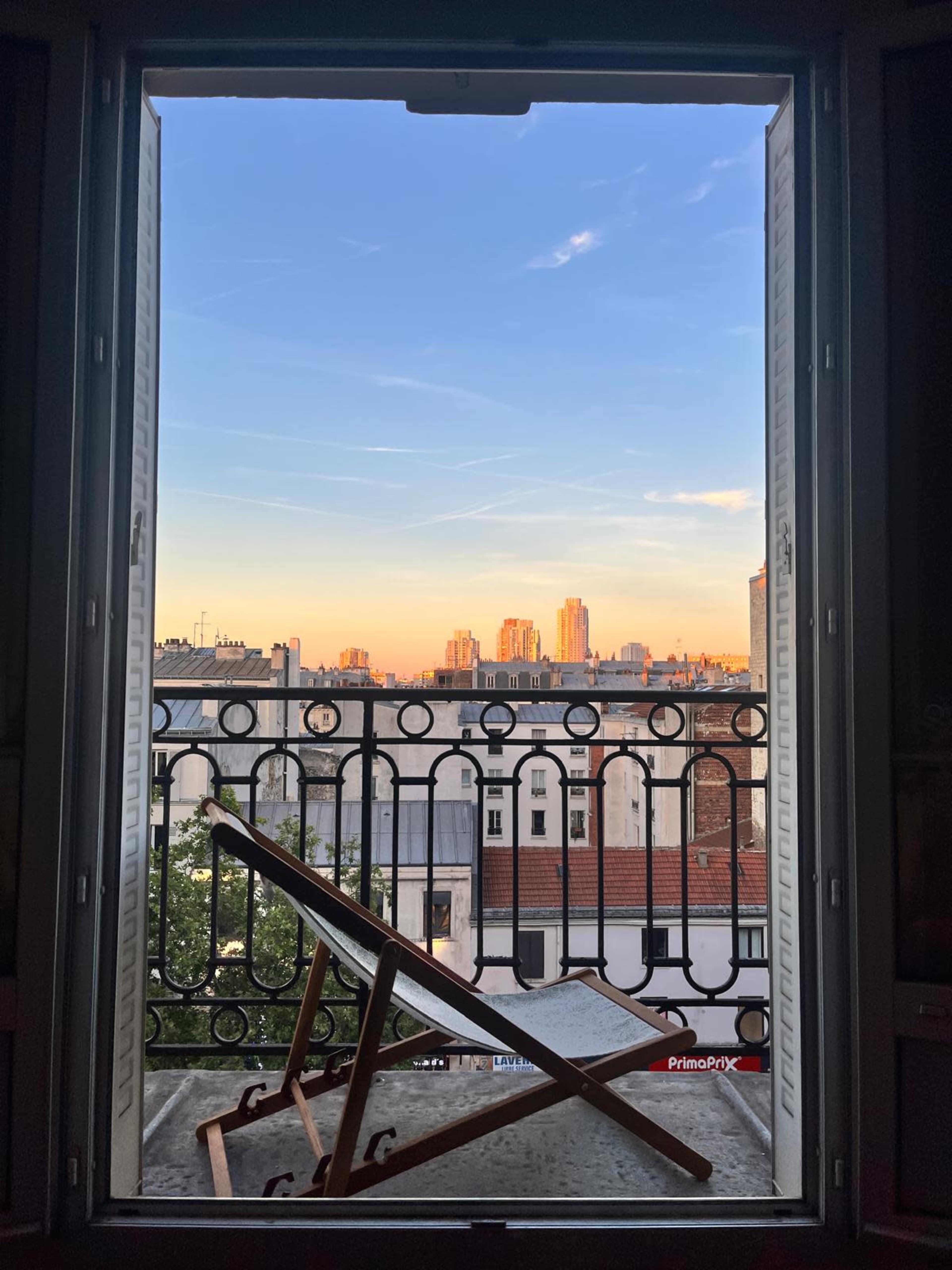 The image shows a balcony window open to a city skyline during sunset, with a wooden lounge chair positioned inside.