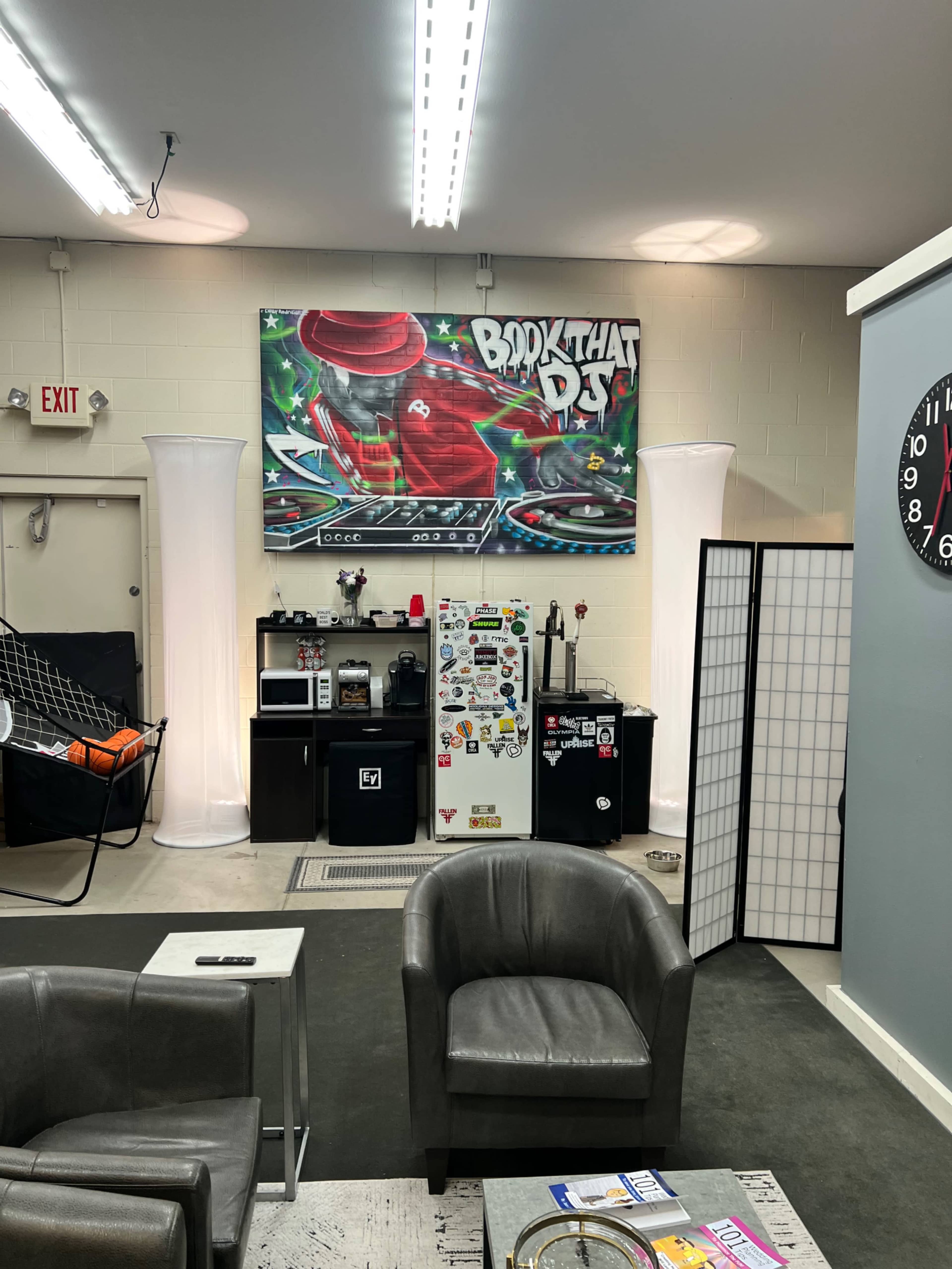 A modern office space with a colorful mural on the wall, featuring a DJ theme, and various furniture including chairs, a coffee table, and a kitchenette area with a fridge and microwave.