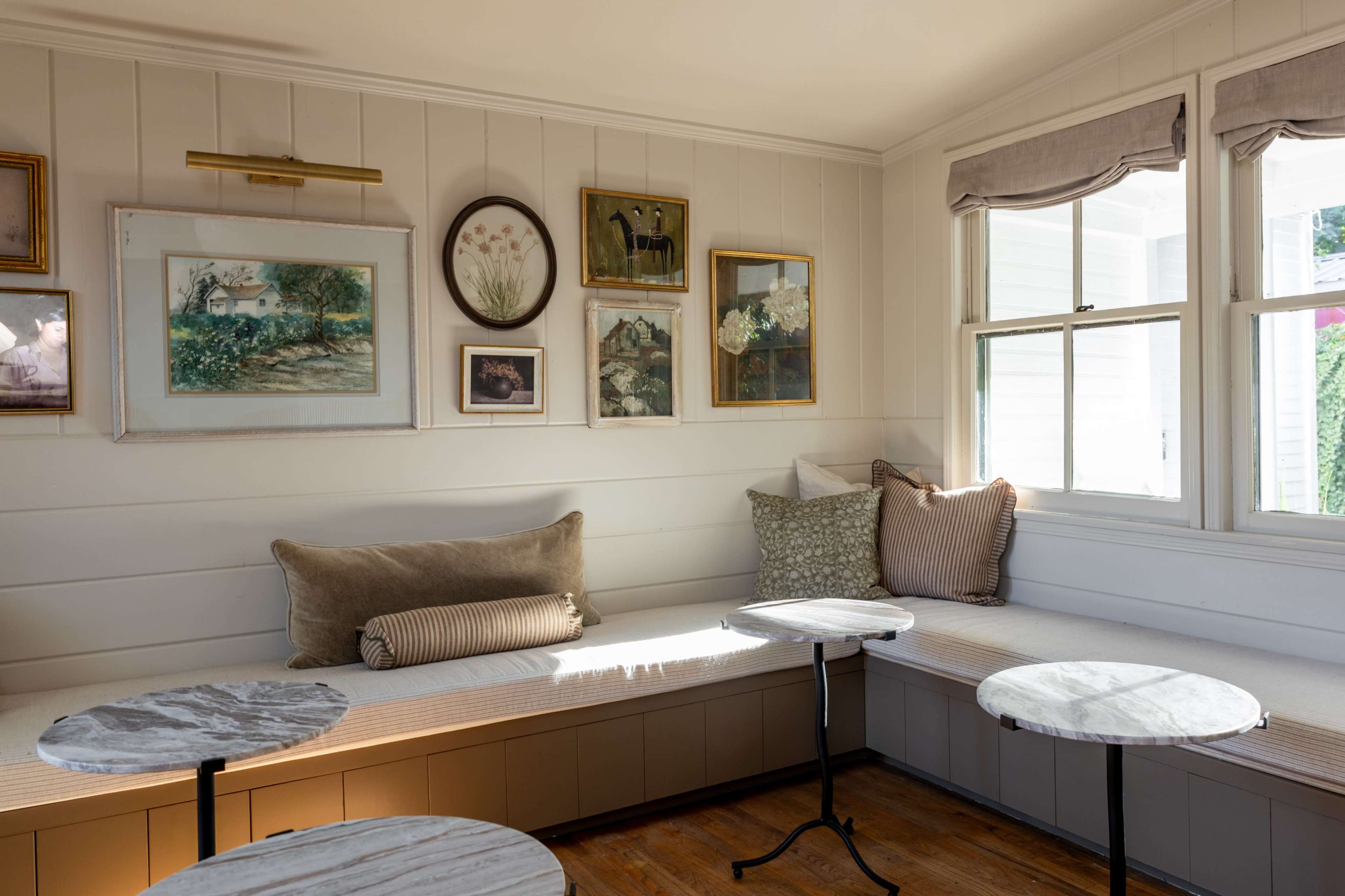 The image shows a cozy corner of a room with a built-in bench, decorative pillows, and framed artwork on the walls, along with small round tables.