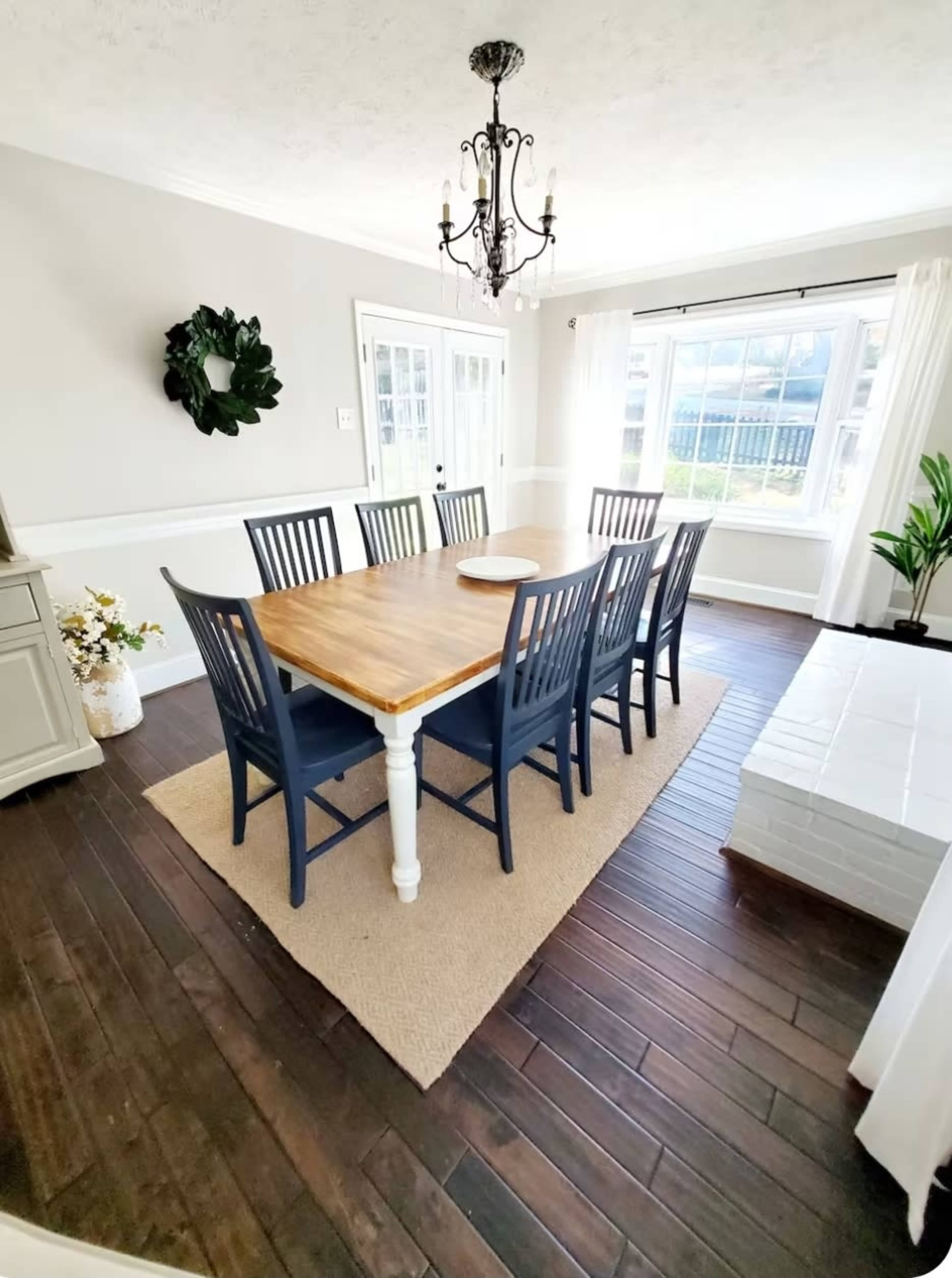 A dining room features a large wooden table surrounded by eight blue chairs, with a chandelier overhead and large windows allowing natural light.