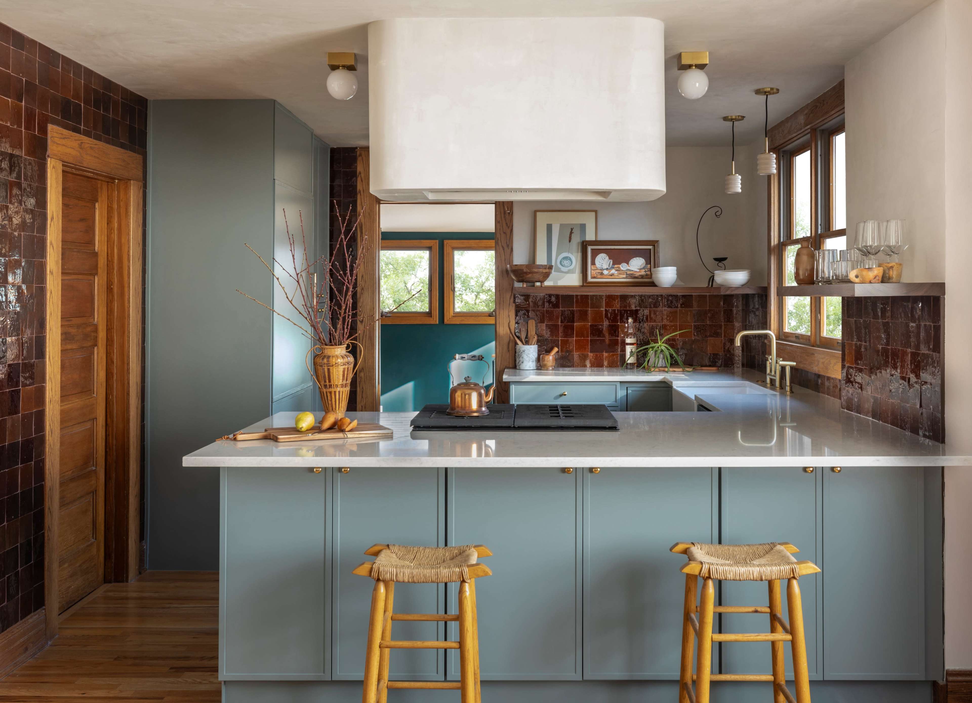 A modern kitchen features a central island with bar stools, green cabinetry, a large range hood, and warm-toned tile accents.