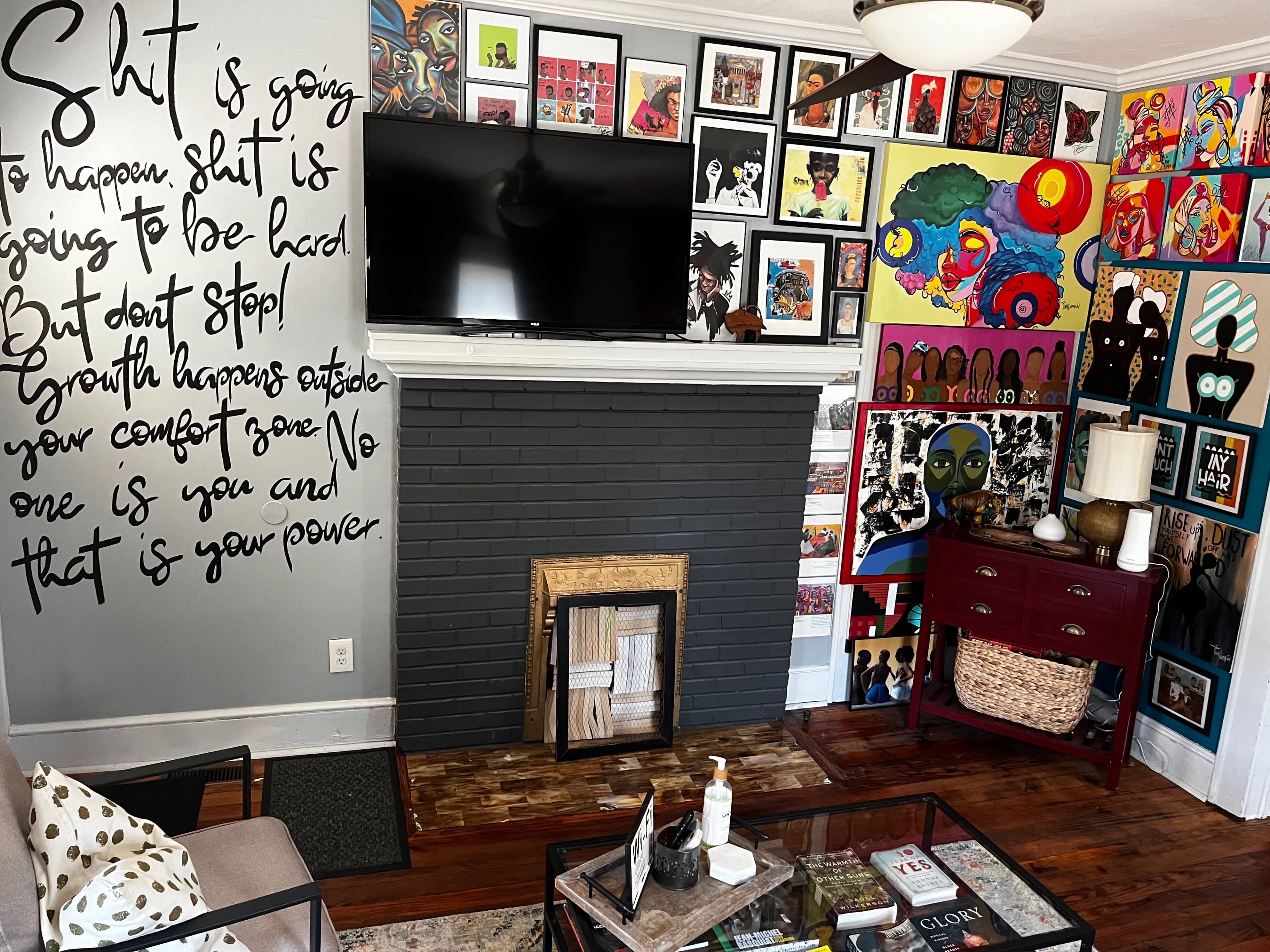 The room features a decorative wall with colorful art, a large television above a dark fireplace, and a coffee table with various items.
