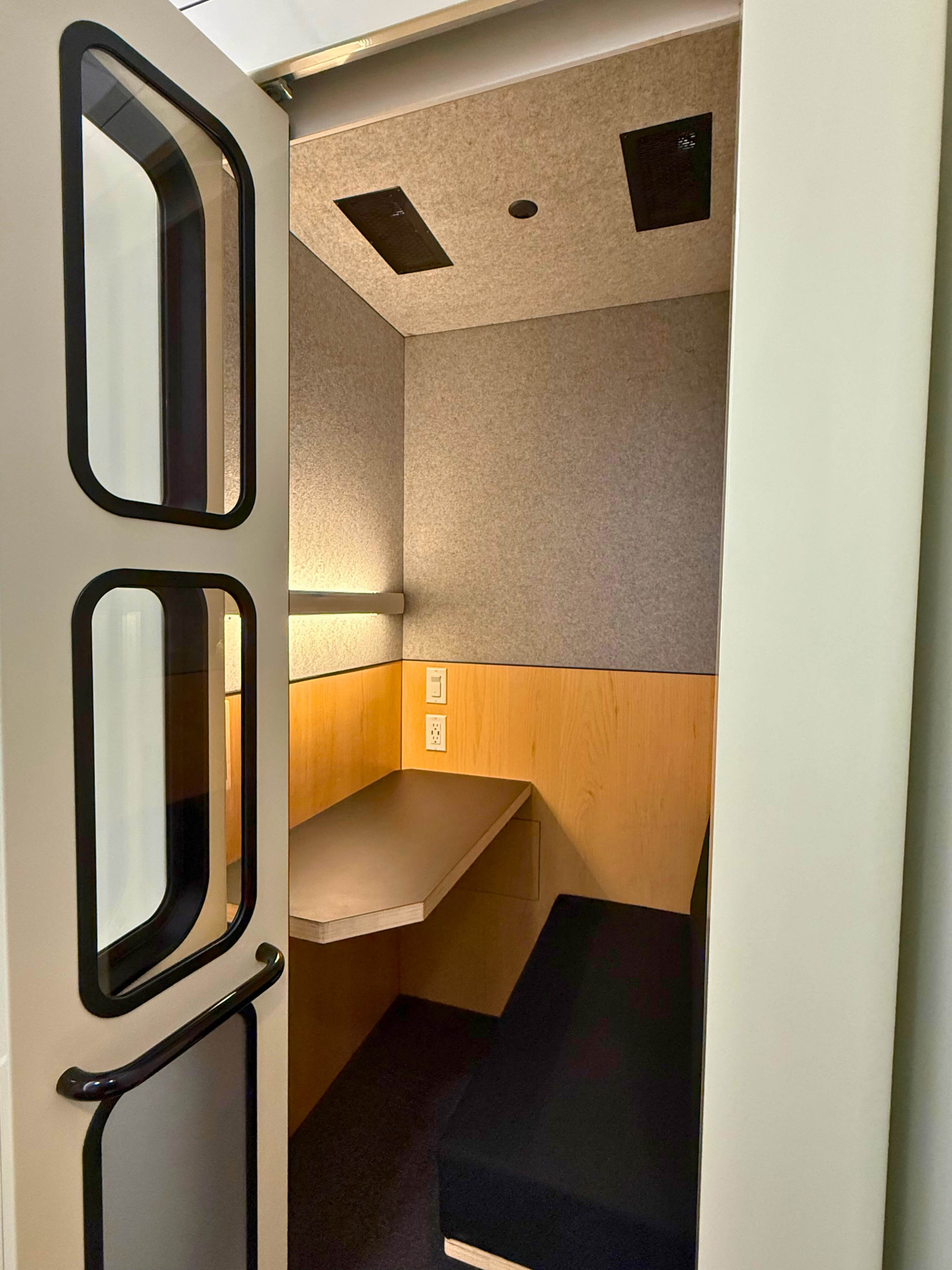 The image shows a small, enclosed space with a bench and a desk, featuring gray walls and a wooden accent, illuminated by a ceiling light.