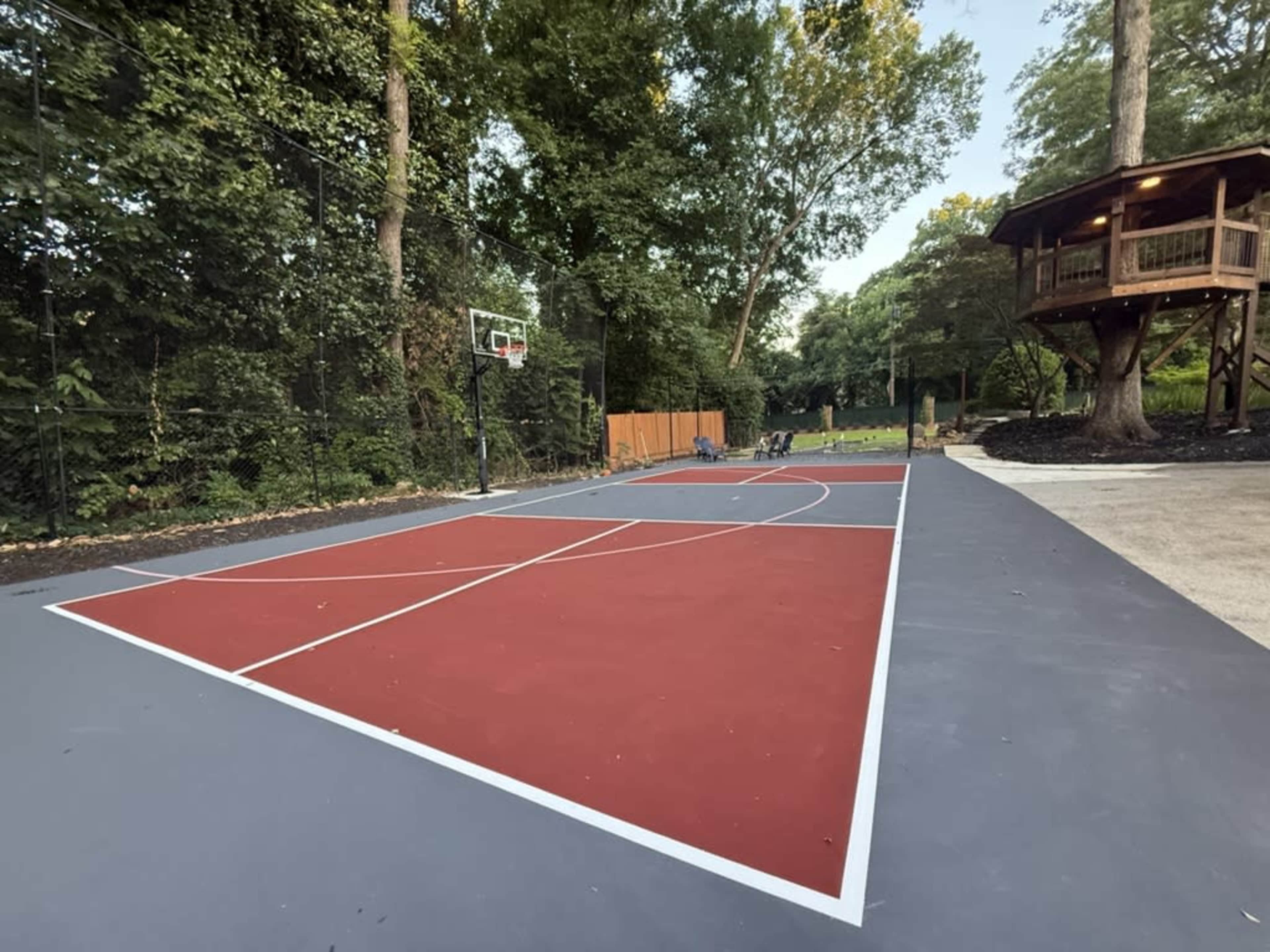 The image shows a basketball court with a red and gray playing surface surrounded by trees and a wooden treehouse in the background.