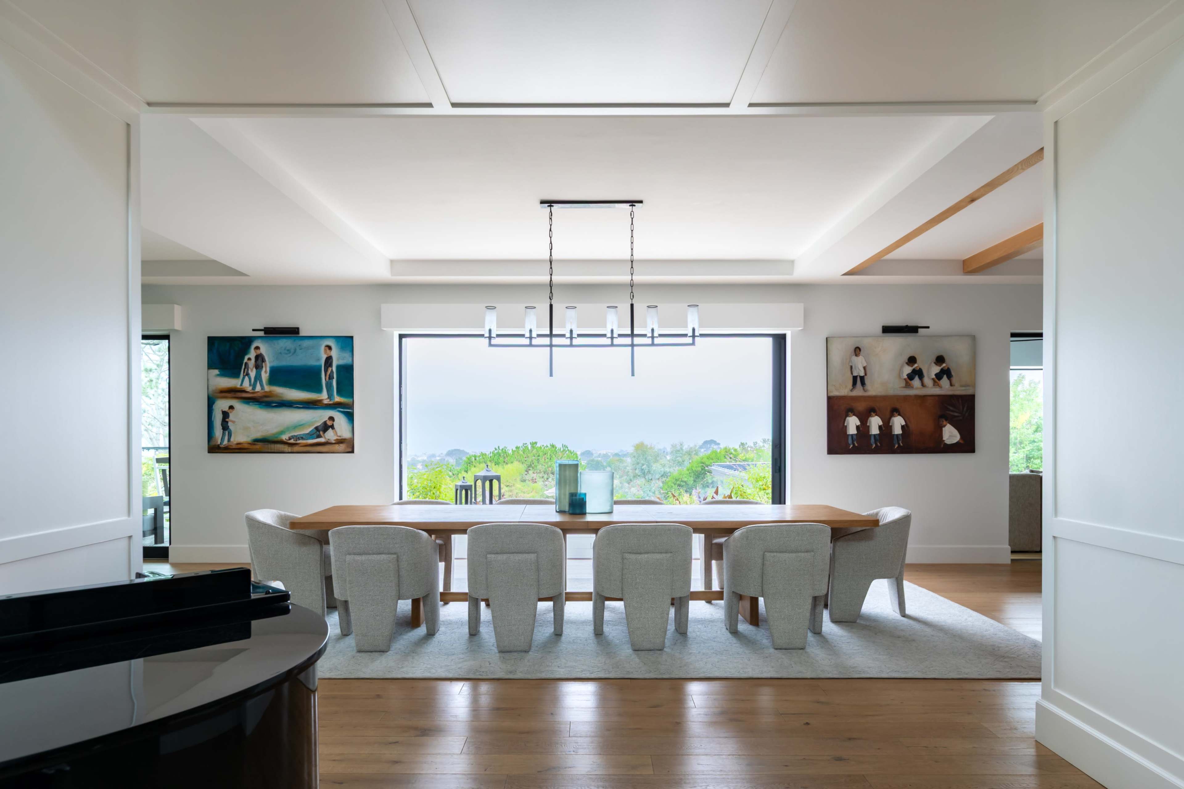 A long dining table with upholstered chairs is set in a bright room featuring large windows and artwork on the walls.