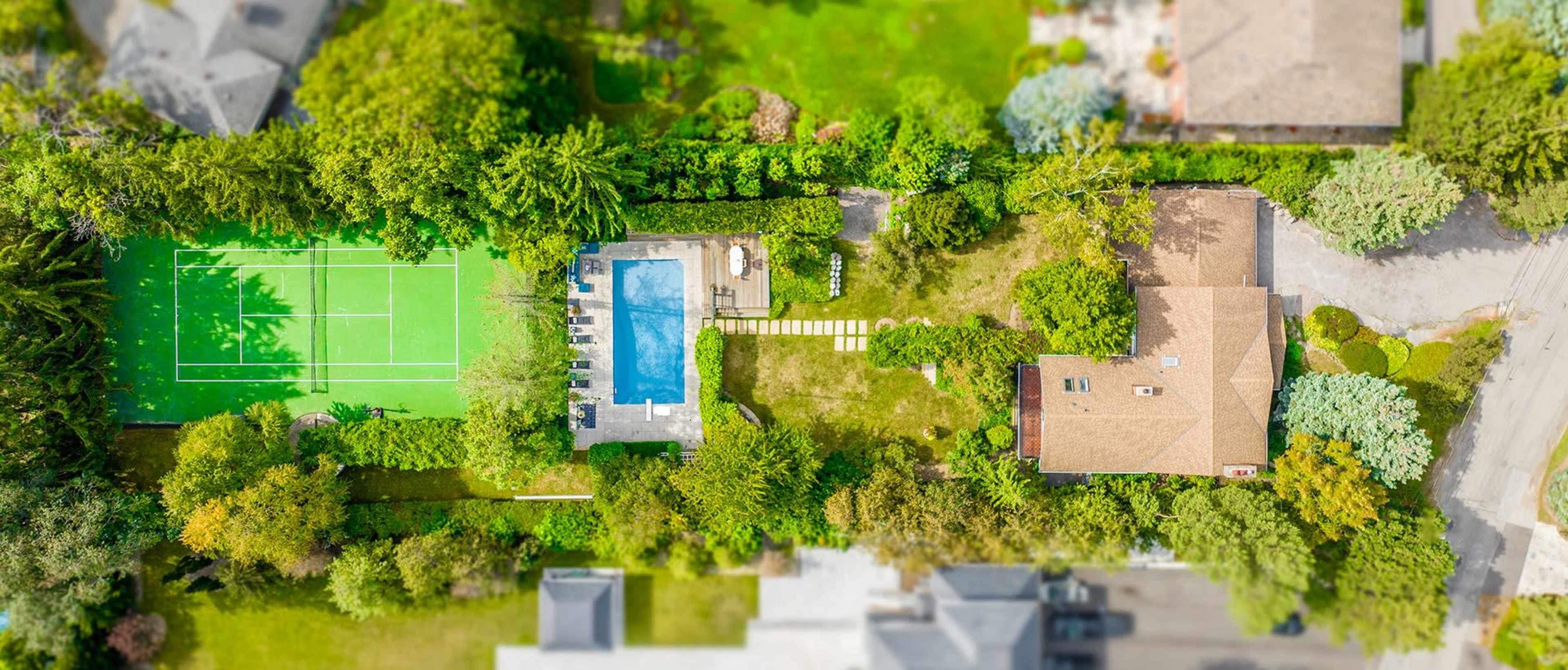 A backyard with a swimming pool and a tennis court, surrounded by dense greenery and a nearby house.