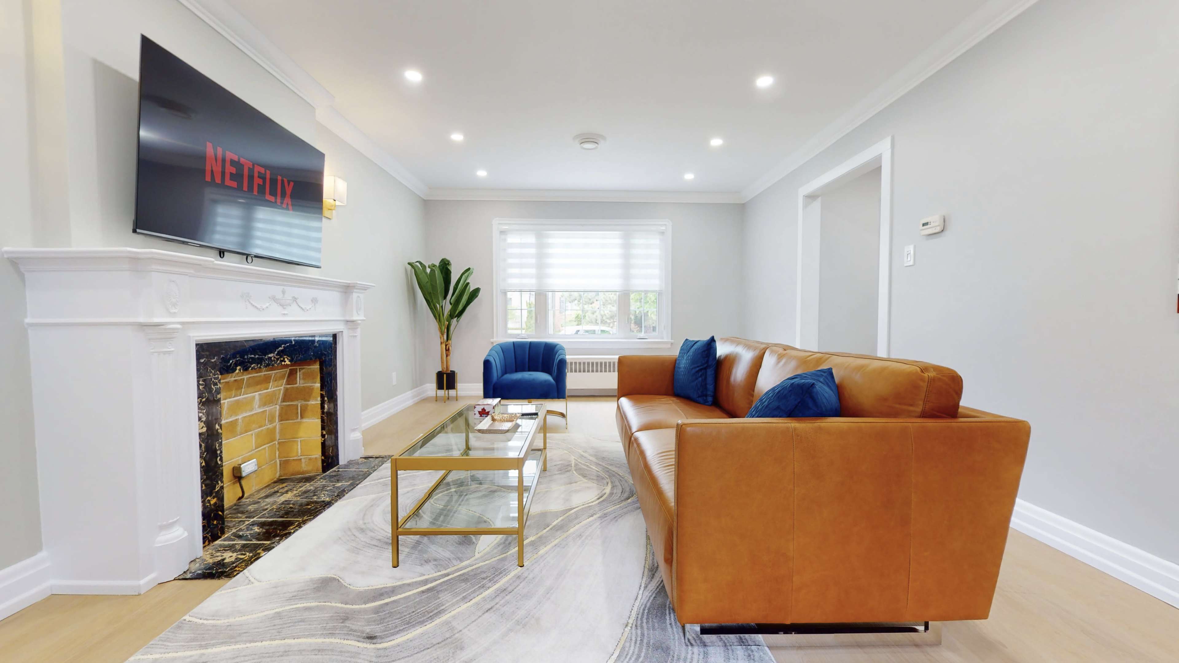A modern living room features a brown leather sofa, a glass coffee table, a fireplace with a yellow brick, and a large TV displaying the Netflix logo.