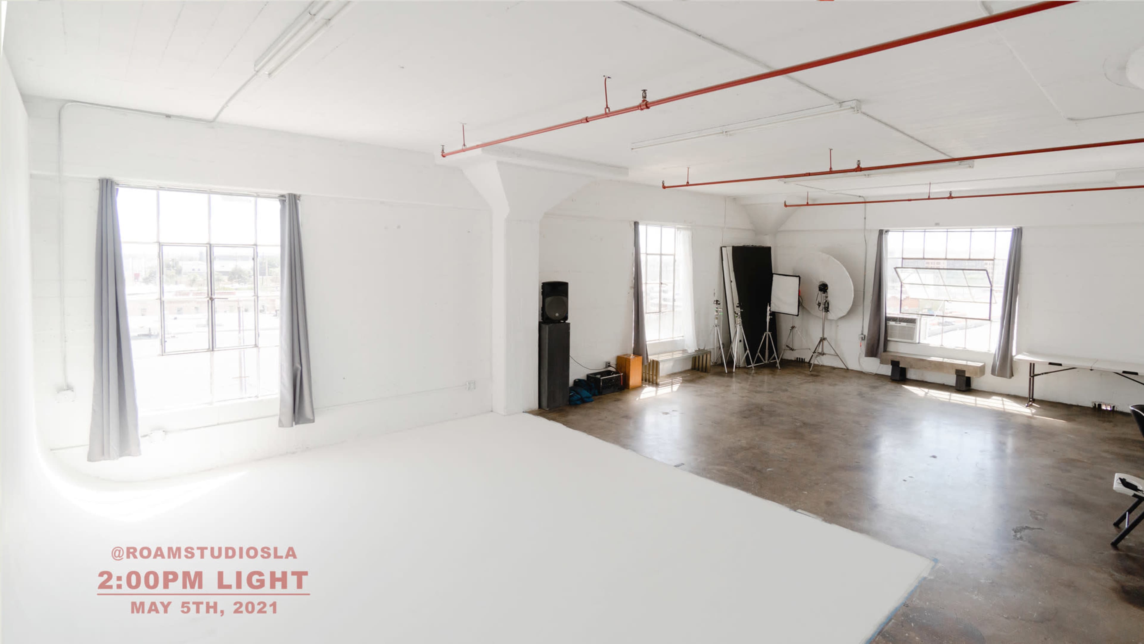 A spacious, well-lit studio with large windows and a simple setup featuring a white backdrop, lighting equipment, and minimal furnishings.
